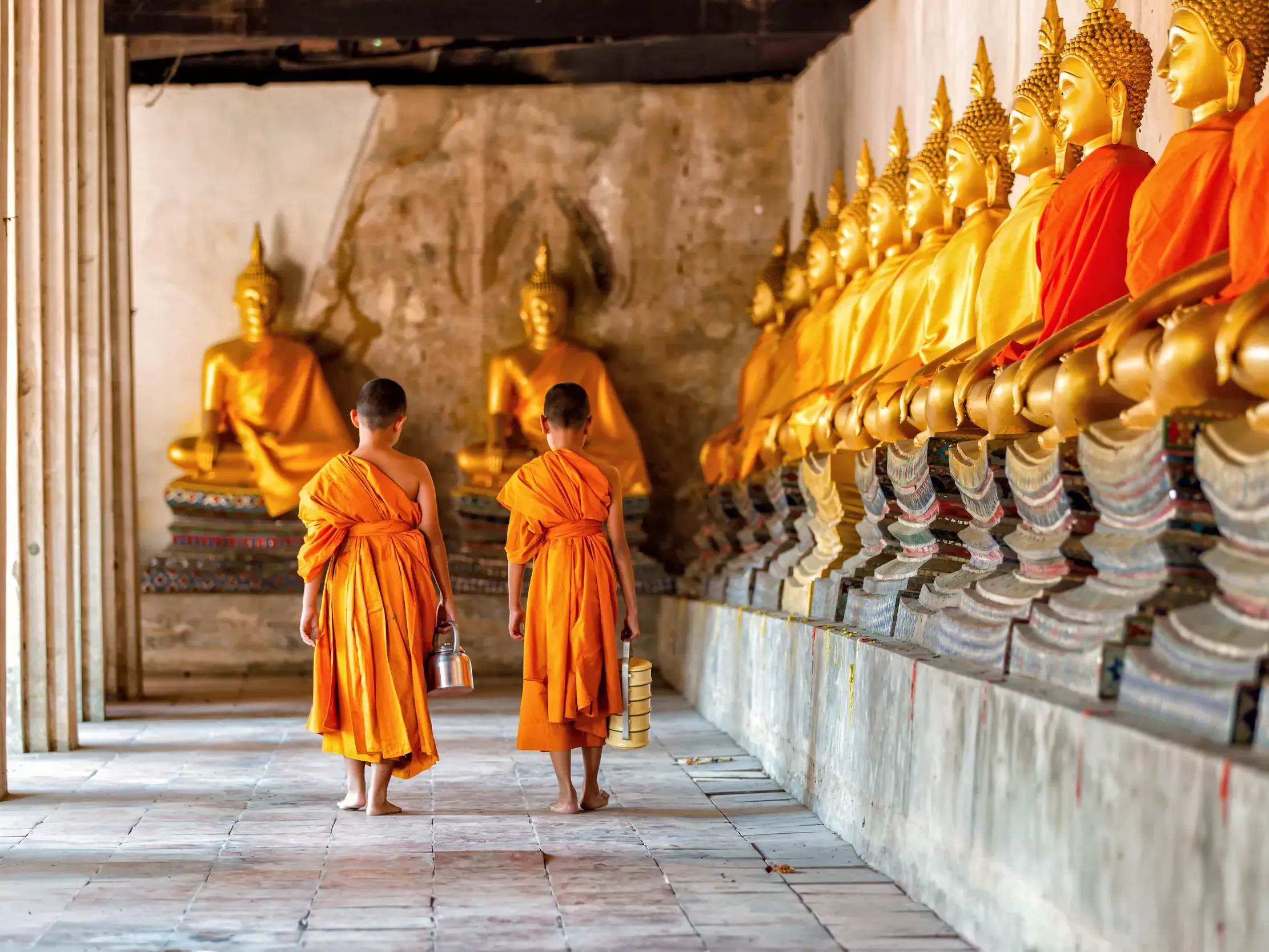 Zwei junge Mönche gehen barfuß durch einen Tempelgang in Ayutthaya, vorbei an einer langen Reihe goldener Buddha-Statuen.