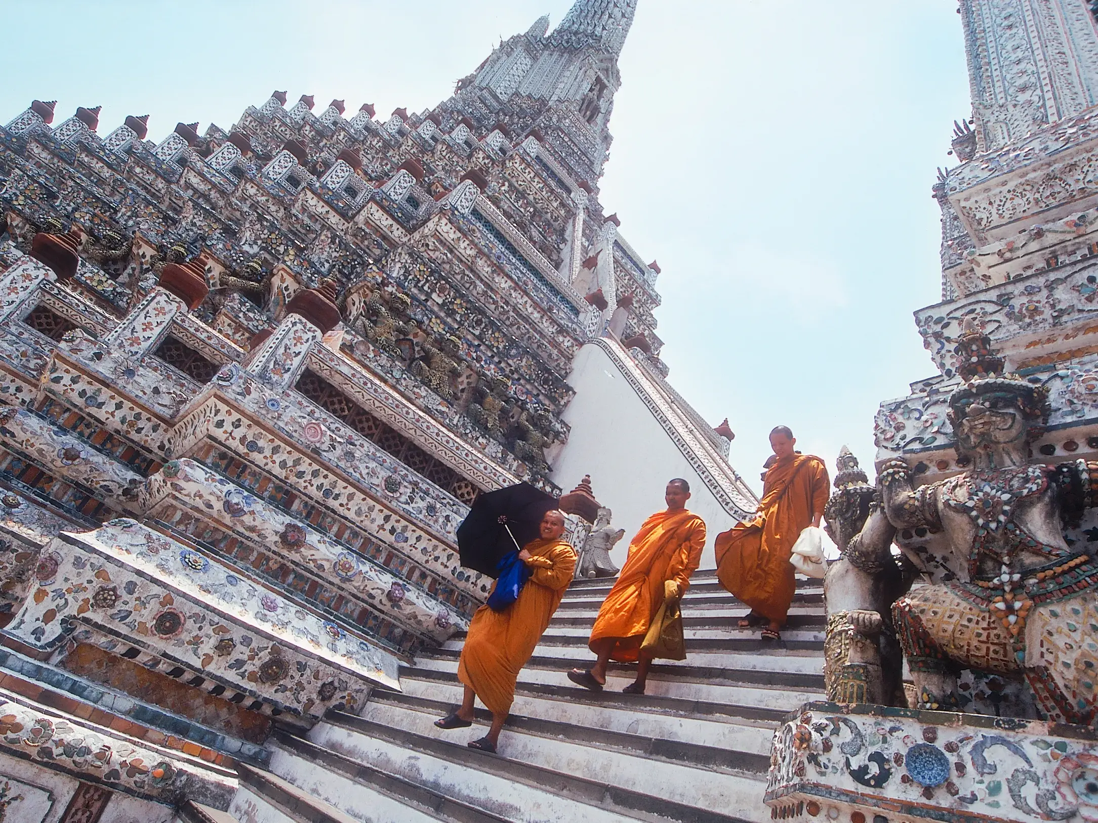 Buddhistische Mönche in orangen Roben steigen die steile, reich verzierte Treppe des Tempels Wat Arun in Bangkok hinab.