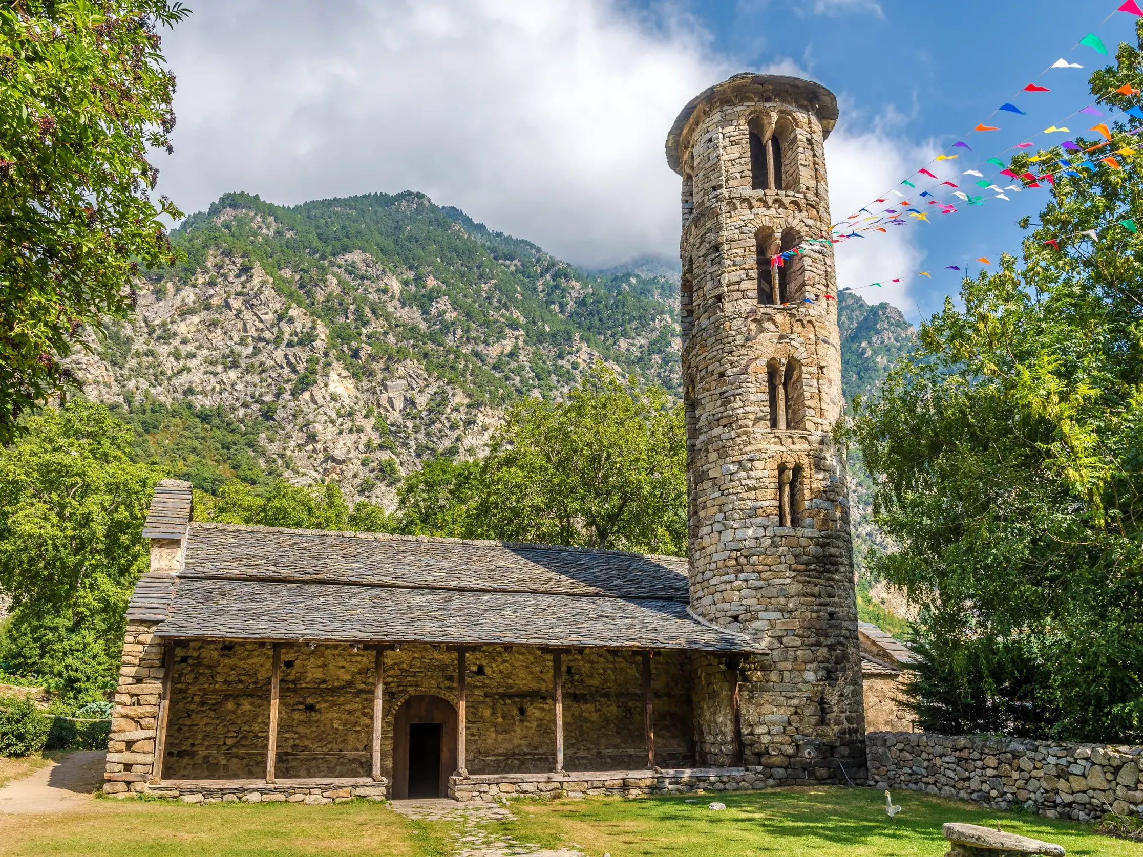 Die romanische Kirche Santa Coloma mit ihrem runden Glockenturm und einer bunten Wimpelkette vor der Bergkulisse der Pyrenäen.