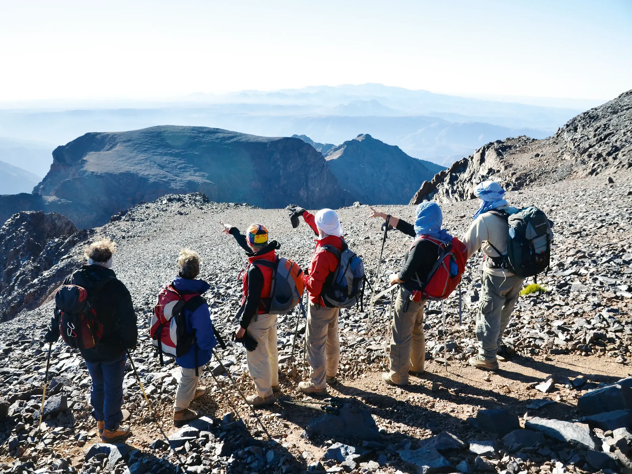 Ein Hauser-Reiseleiter zeigt seiner Trekkinggruppe auf einem steinigen Bergpfad im Hohen Atlas die weite Gipfellandschaft.