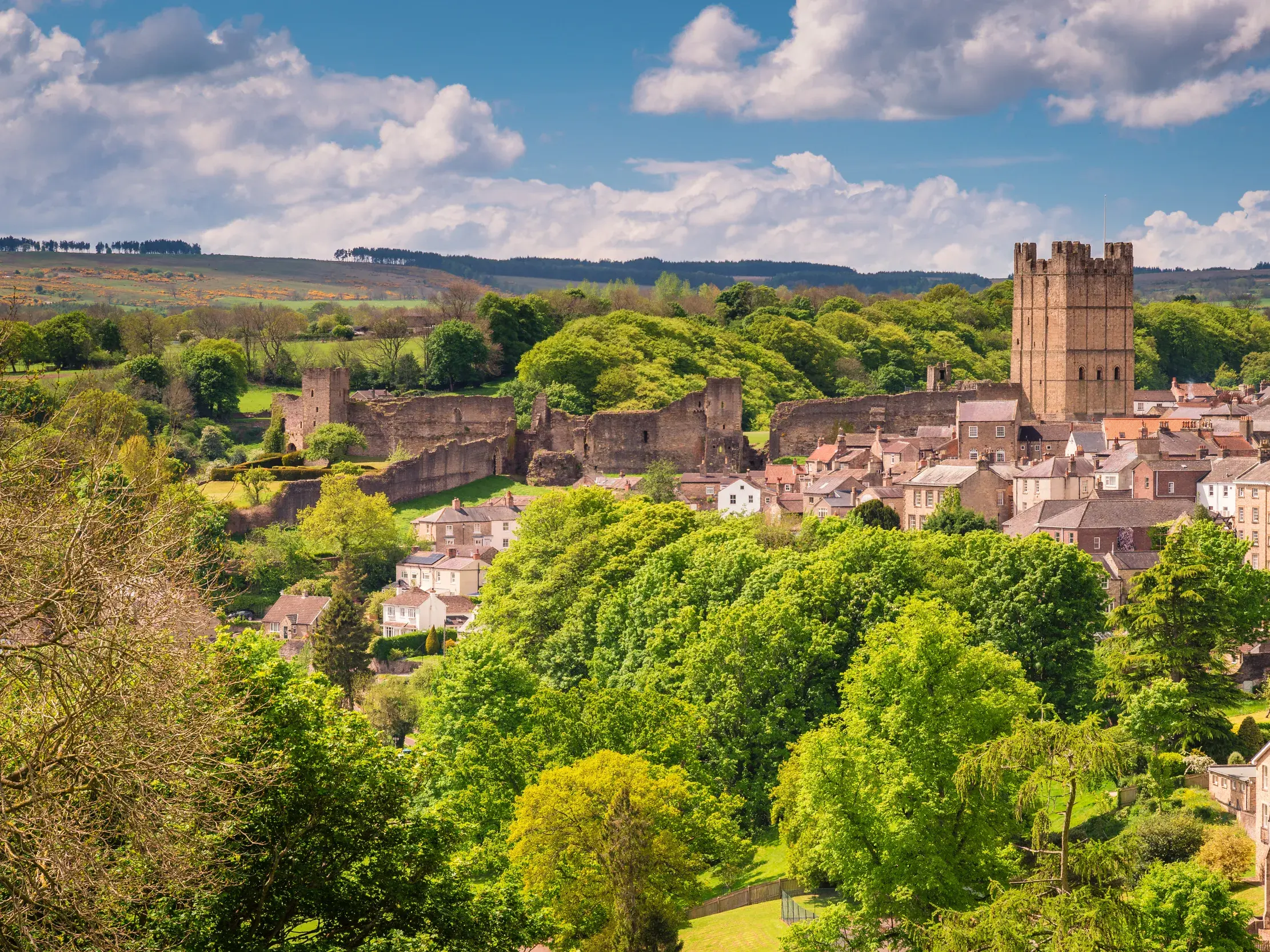 Blick auf die historische Stadt Richmond in Yorkshire mit dem Richmond Castle, umgeben von grünen Hügeln an einem sonnigen Tag.