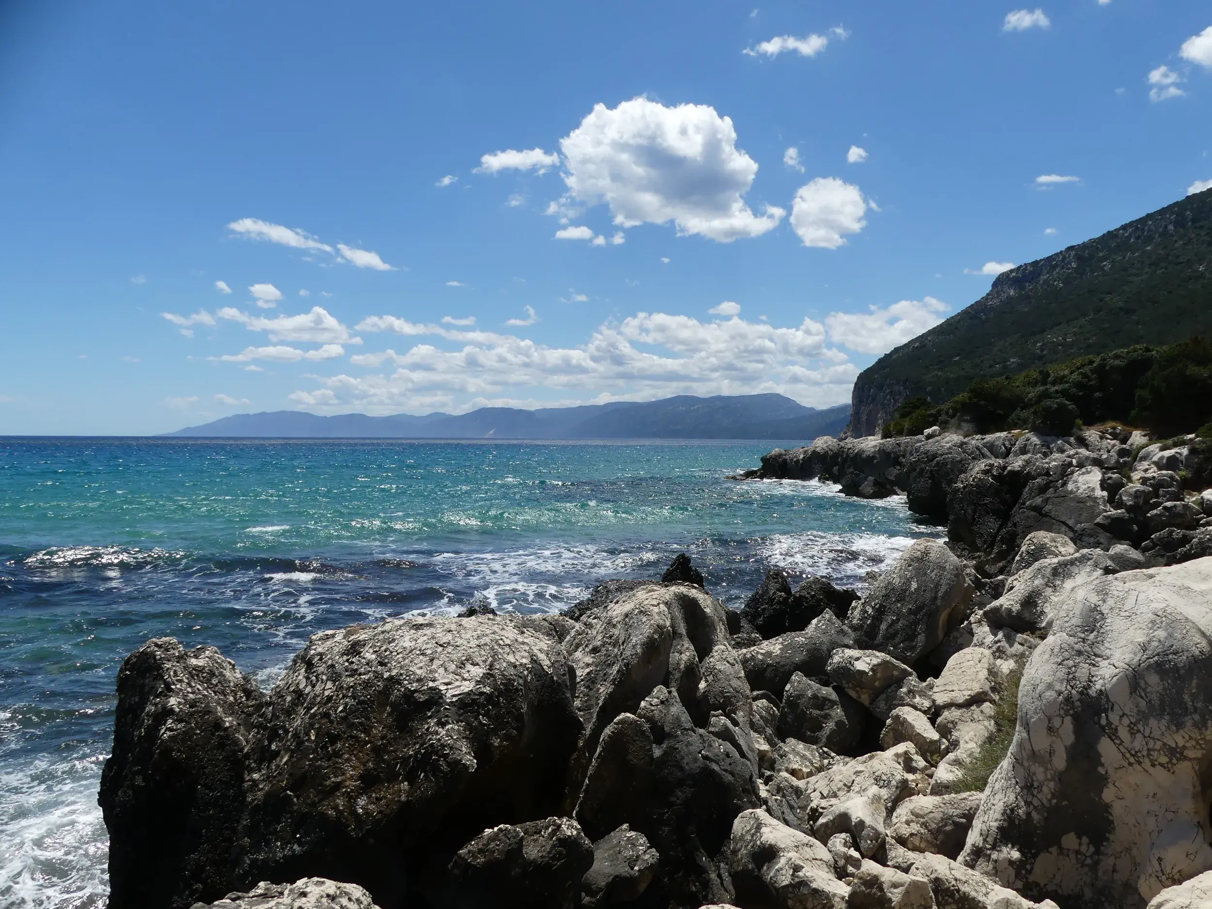 Blick von den Felsen auf die wilde Küste Sardiniens mit türkisblauem Meer und bewachsenen Steilhängen.