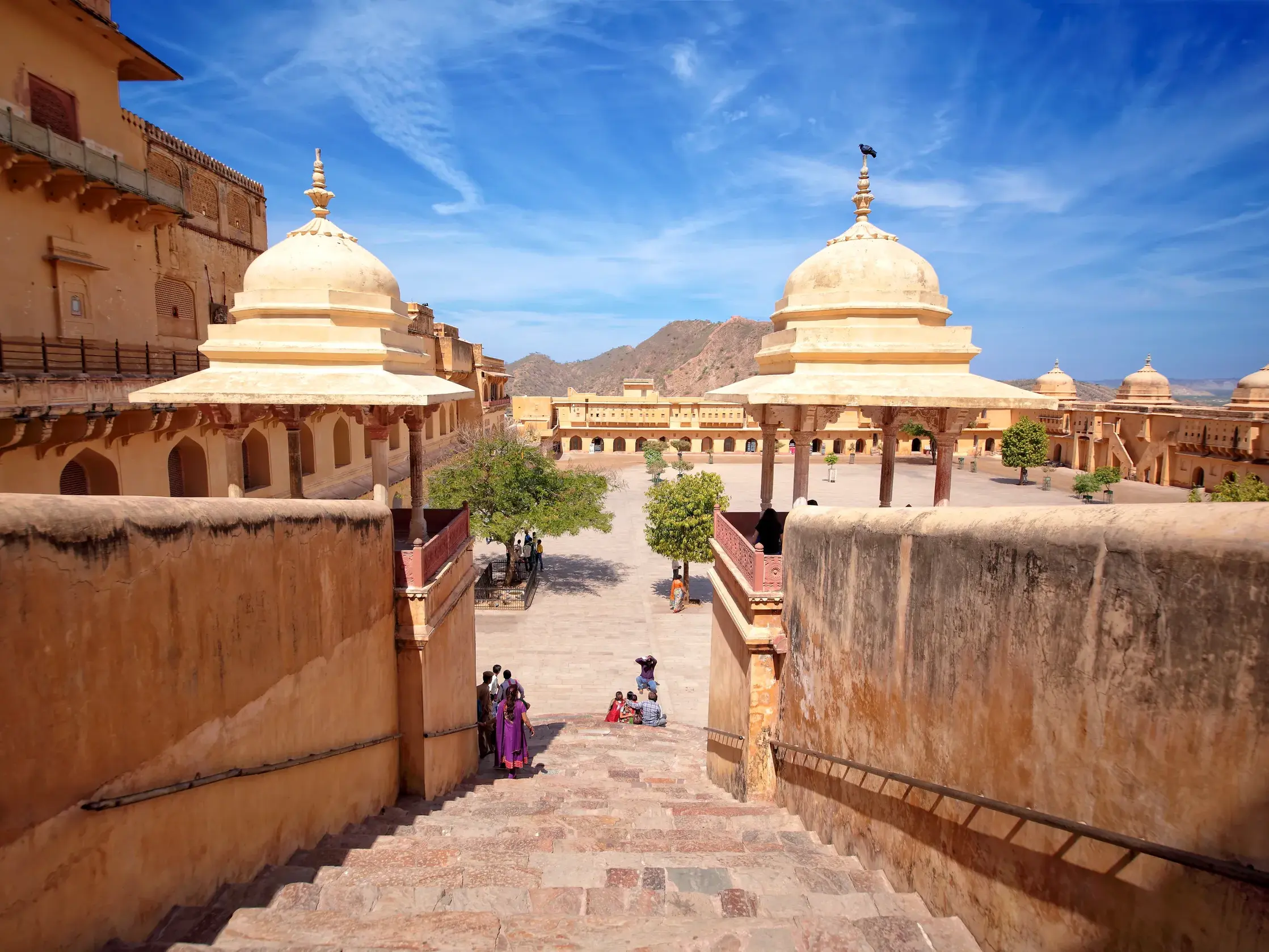 Blick von einer Steintreppe auf den sonnigen Innenhof der Festung von Amber in Jaipur mit Kuppelpavillons und Besuchern.