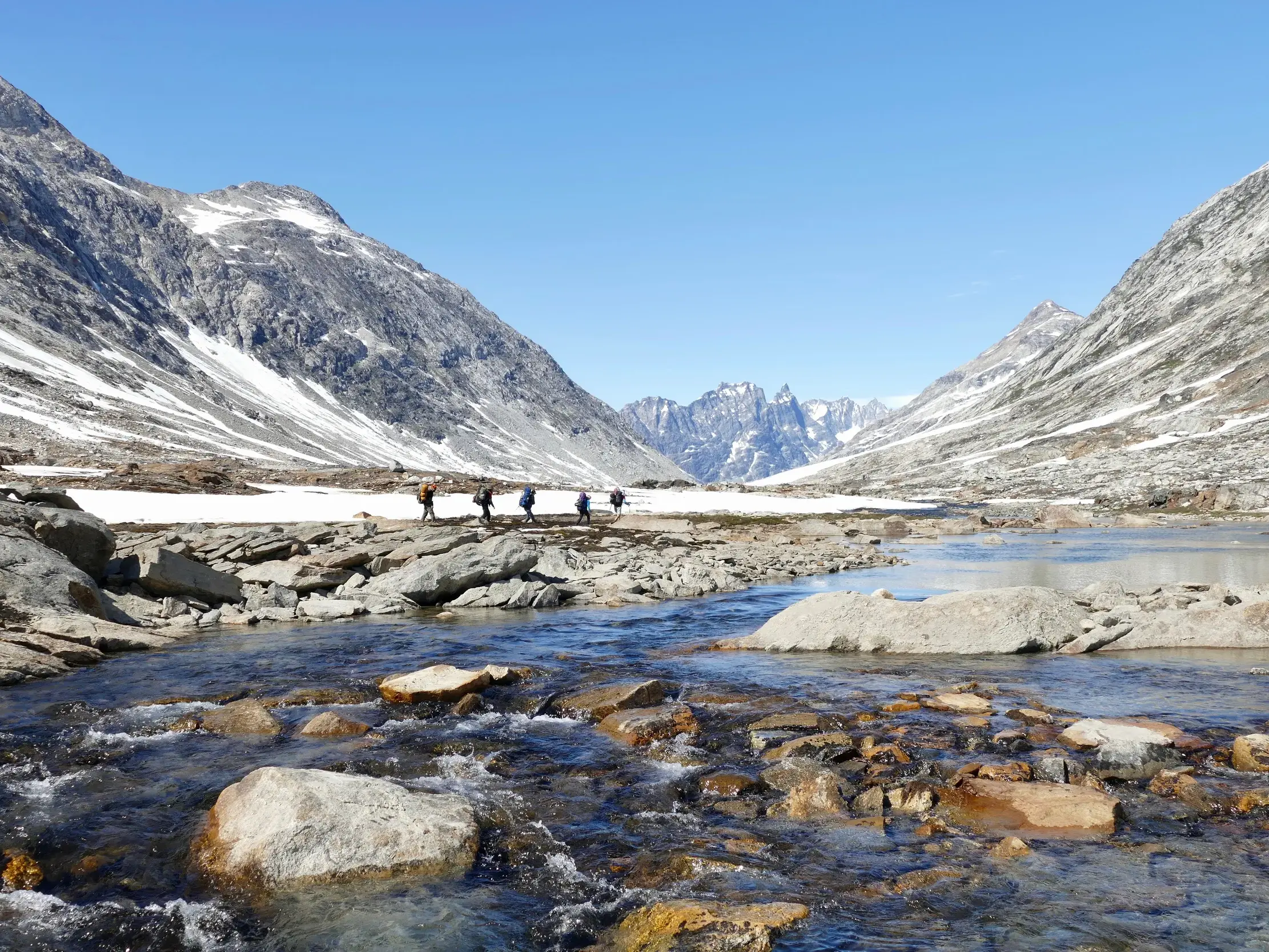 Wanderer mit Rucksäcken durchqueren ein arktisches Tal in Ostgrönland, flankiert von schneebedeckten Bergen und einem klaren Fluss.