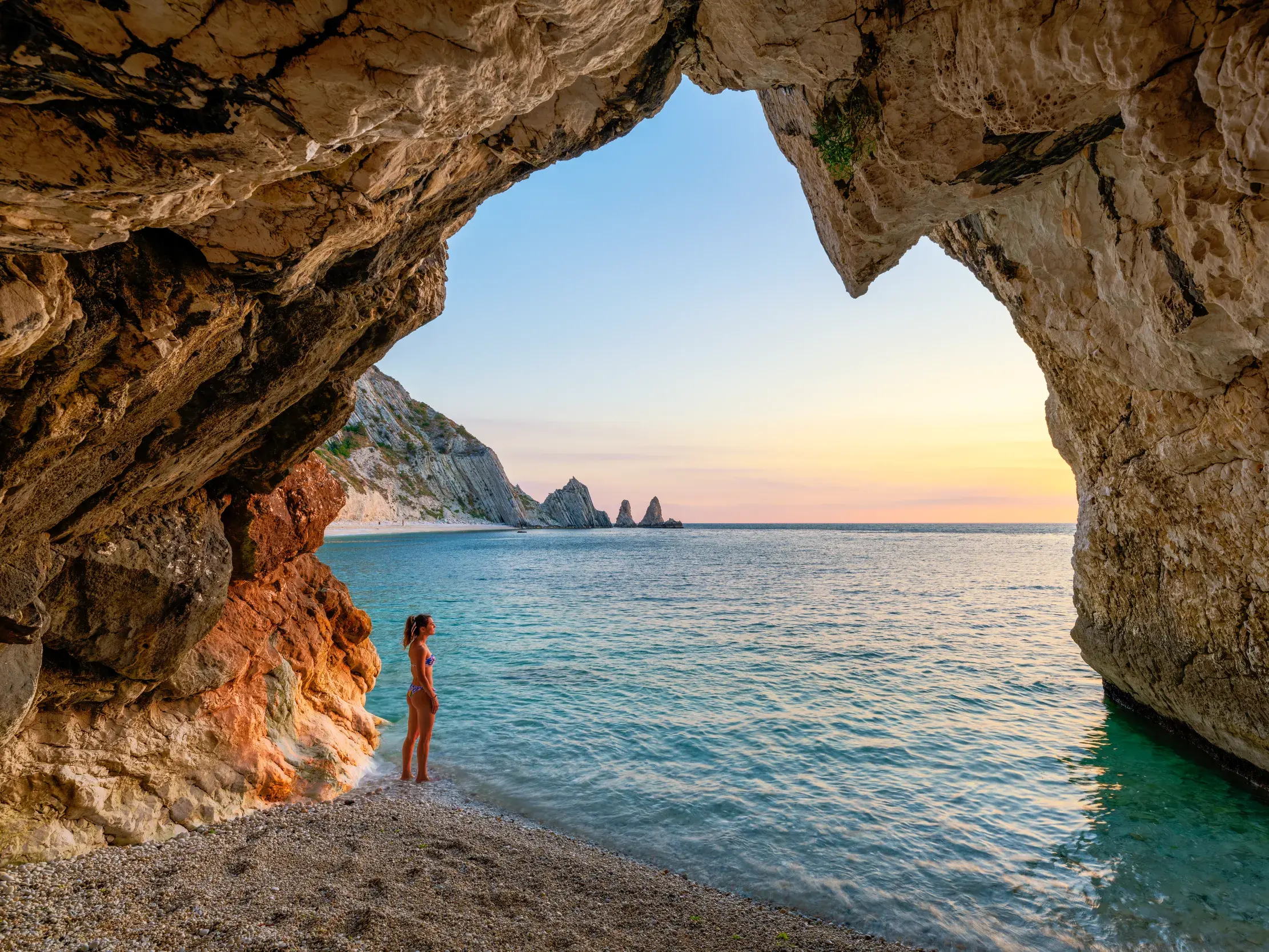 Blick aus einer Felsenhöhle auf die Steilküste des Monte Conero mit dem türkisblauen Meer der Adria bei Sonnenuntergang.