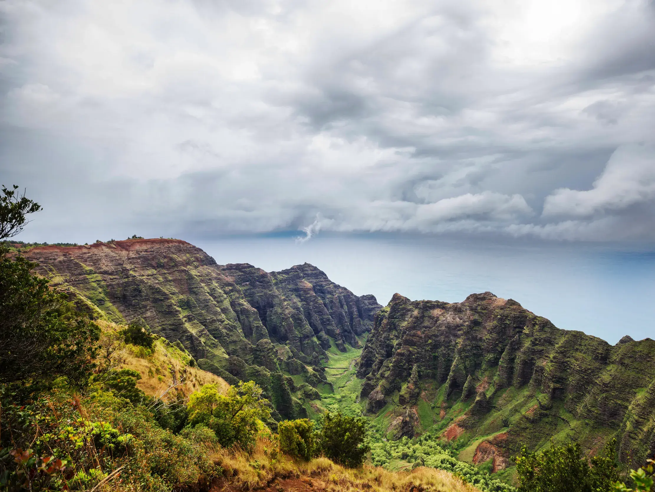 Panoramablick über das grüne Kalalau-Tal an der Nā Pali Küste auf Kauai mit dem Pazifik im Hintergrund unter dramatischen Wolken.