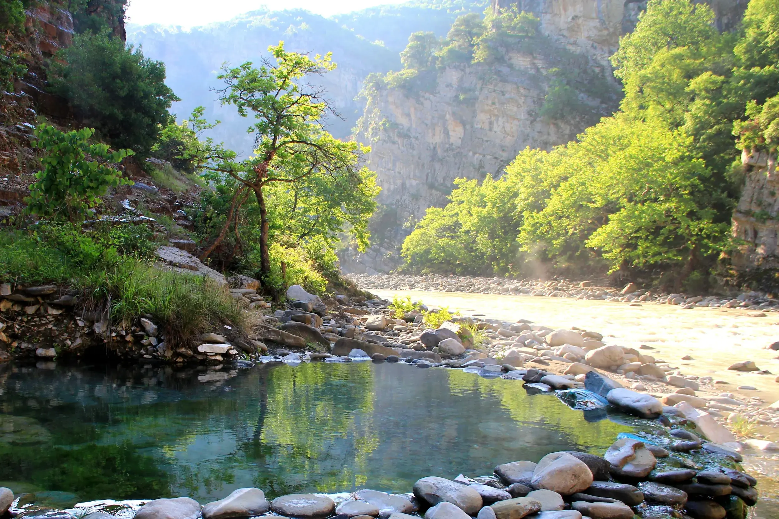 Natürlicher, steingefasster Pool der Thermalquellen von Benjë neben einem Fluss in einer sonnigen Schlucht in Albanien.
