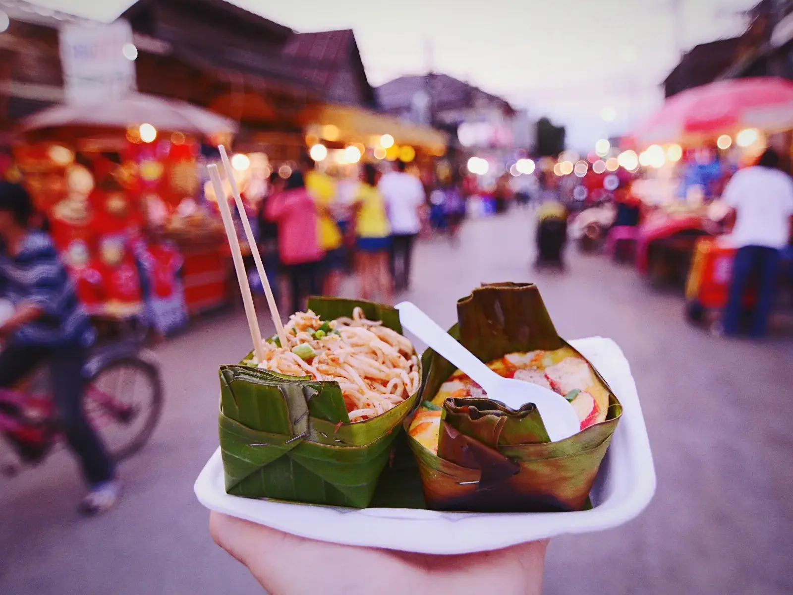 Authentisches Thai-Streetfood in Bananenblatt-Schalen auf einem belebten Nachtmarkt mit leuchtenden Garküchen.