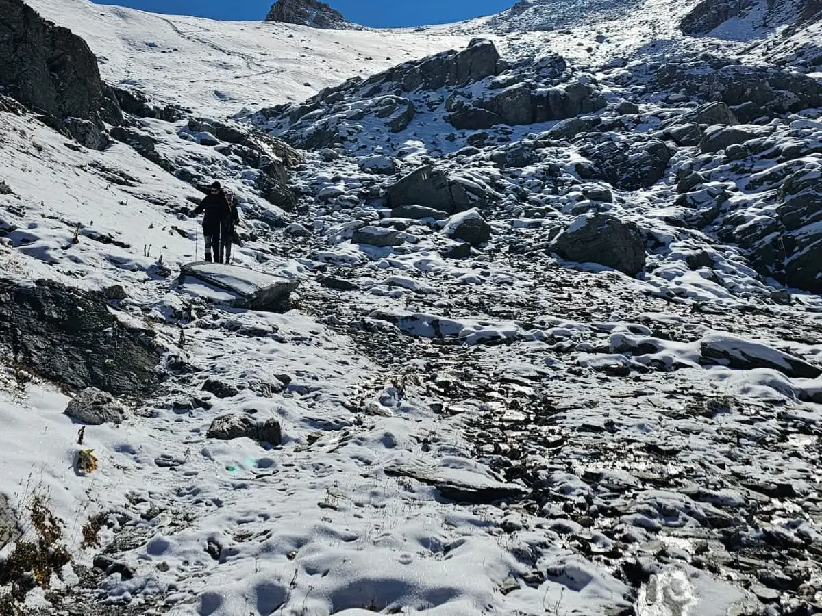 Zwei Wanderer mit Stöcken steigen auf einem schmalen Pfad durch ein schneebedecktes Geröllfeld in den Graubündner Alpen auf.