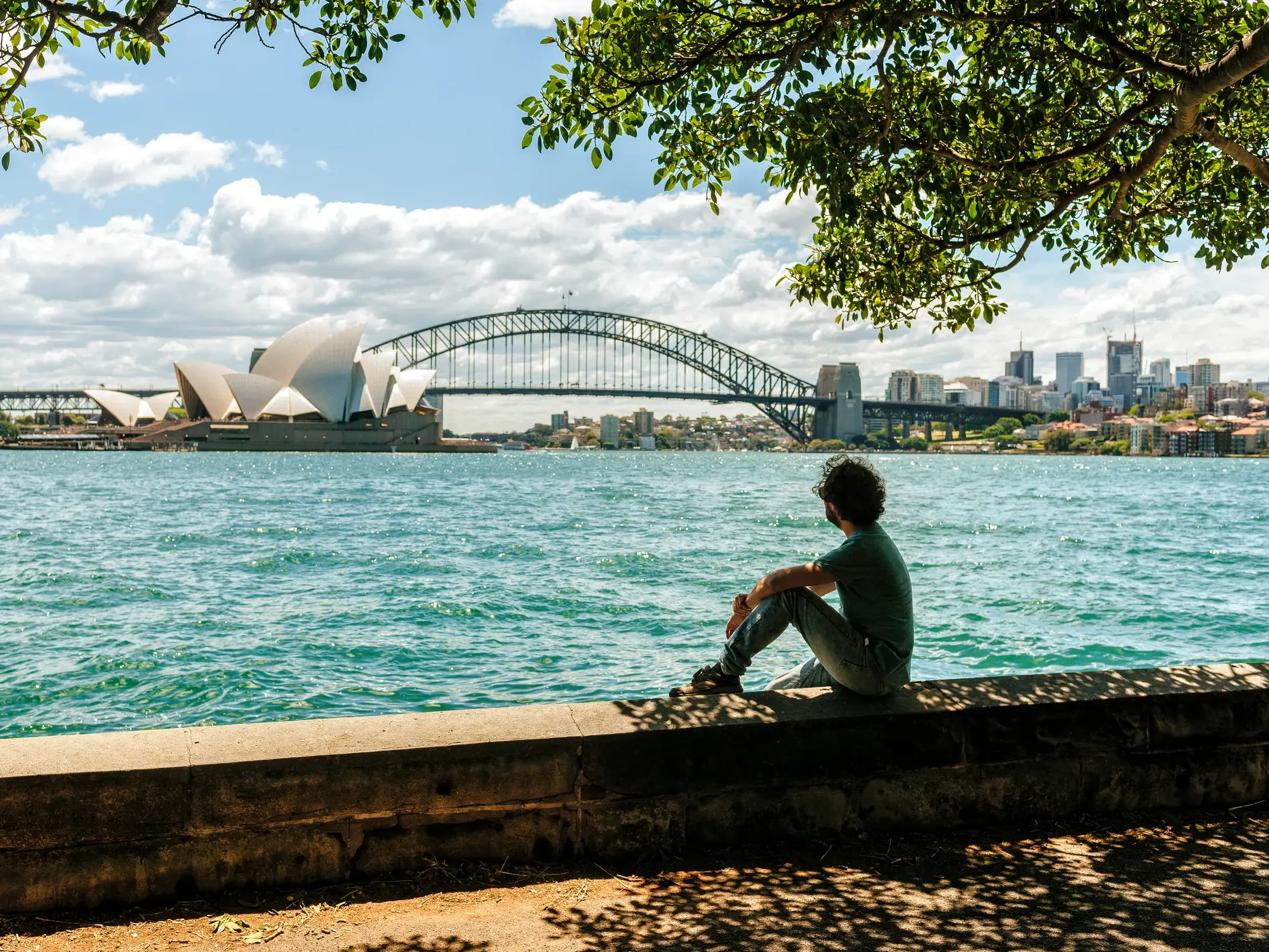 Ein Mann sitzt auf einer Ufermauer in Sydney und blickt nachdenklich auf das Opernhaus und die Harbour Bridge.