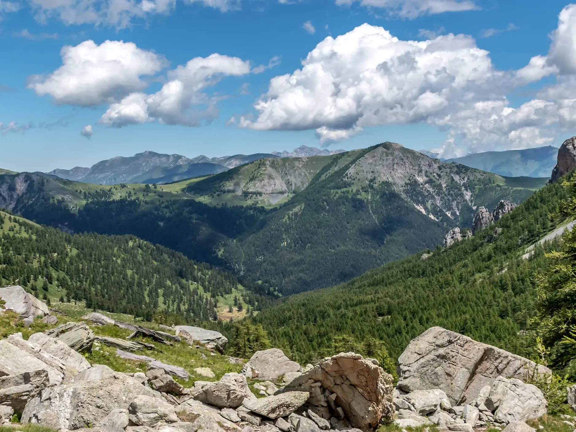 Blick von einem felsigen Wanderweg auf die weite Berglandschaft der Seealpen mit grünen Tälern unter blauem Himmel.