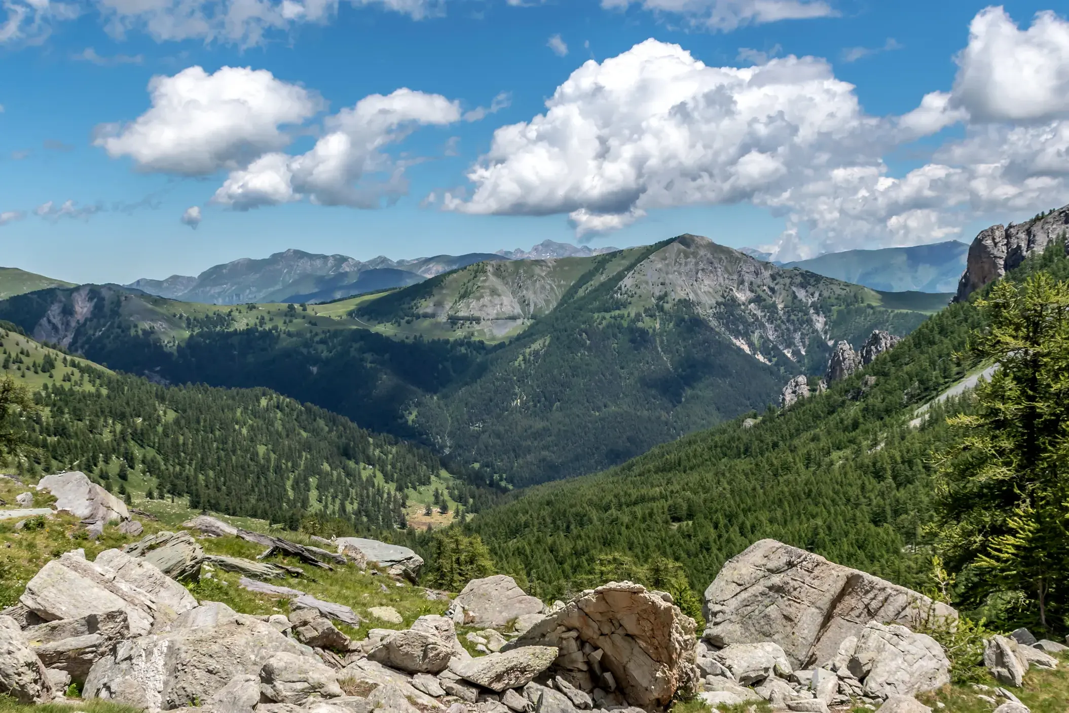 Blick von einem felsigen Wanderweg auf die weite Berglandschaft der Seealpen mit grünen Tälern unter blauem Himmel.