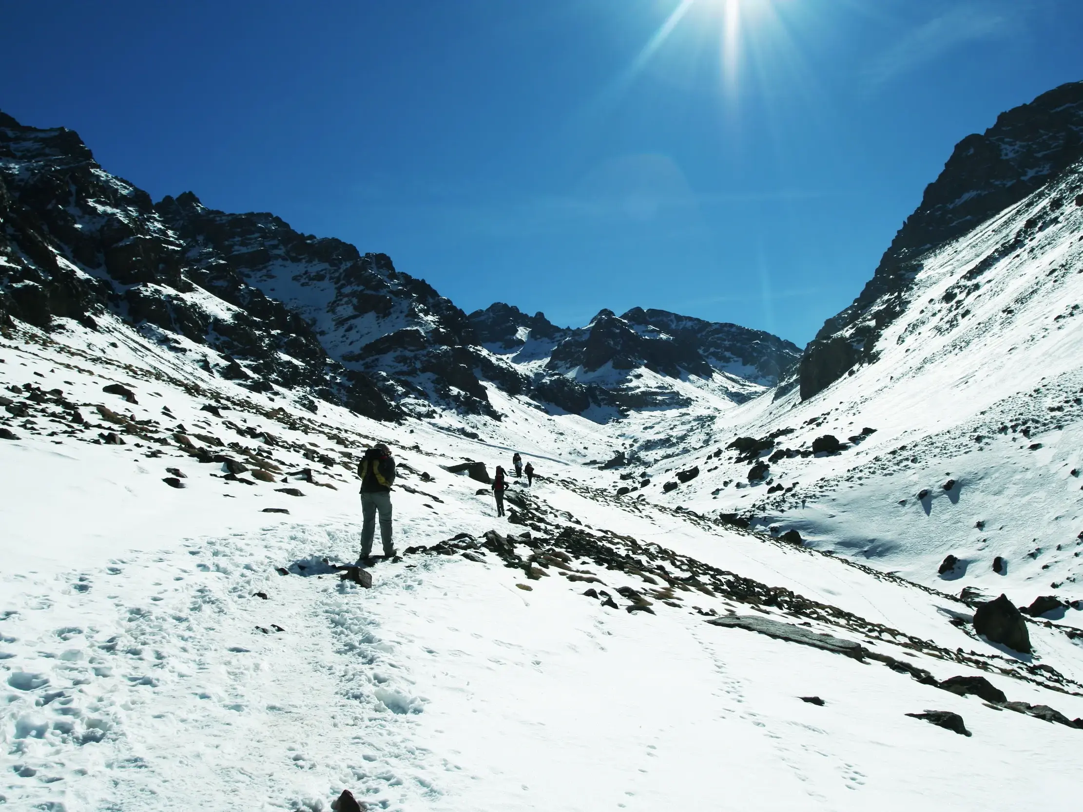 Eine kleine Trekkinggruppe wandert durch ein sonniges, verschneites Tal im Hohen Atlas auf dem Weg zum Gipfel des Toubkal.