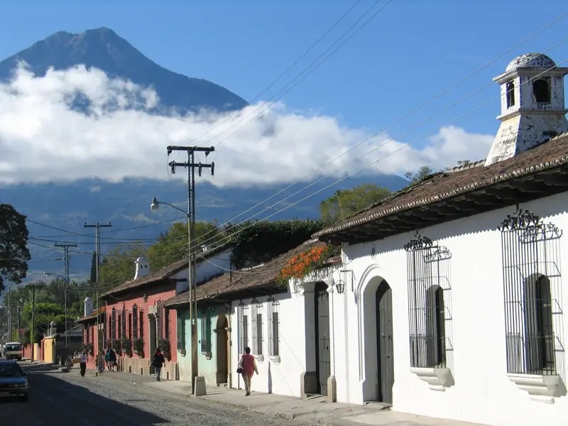 Kopfsteingasse mit bunten Kolonialhäusern in Antigua Guatemala. Im Hintergrund ragt der majestätische Vulkan Agua in den blauen Himmel.