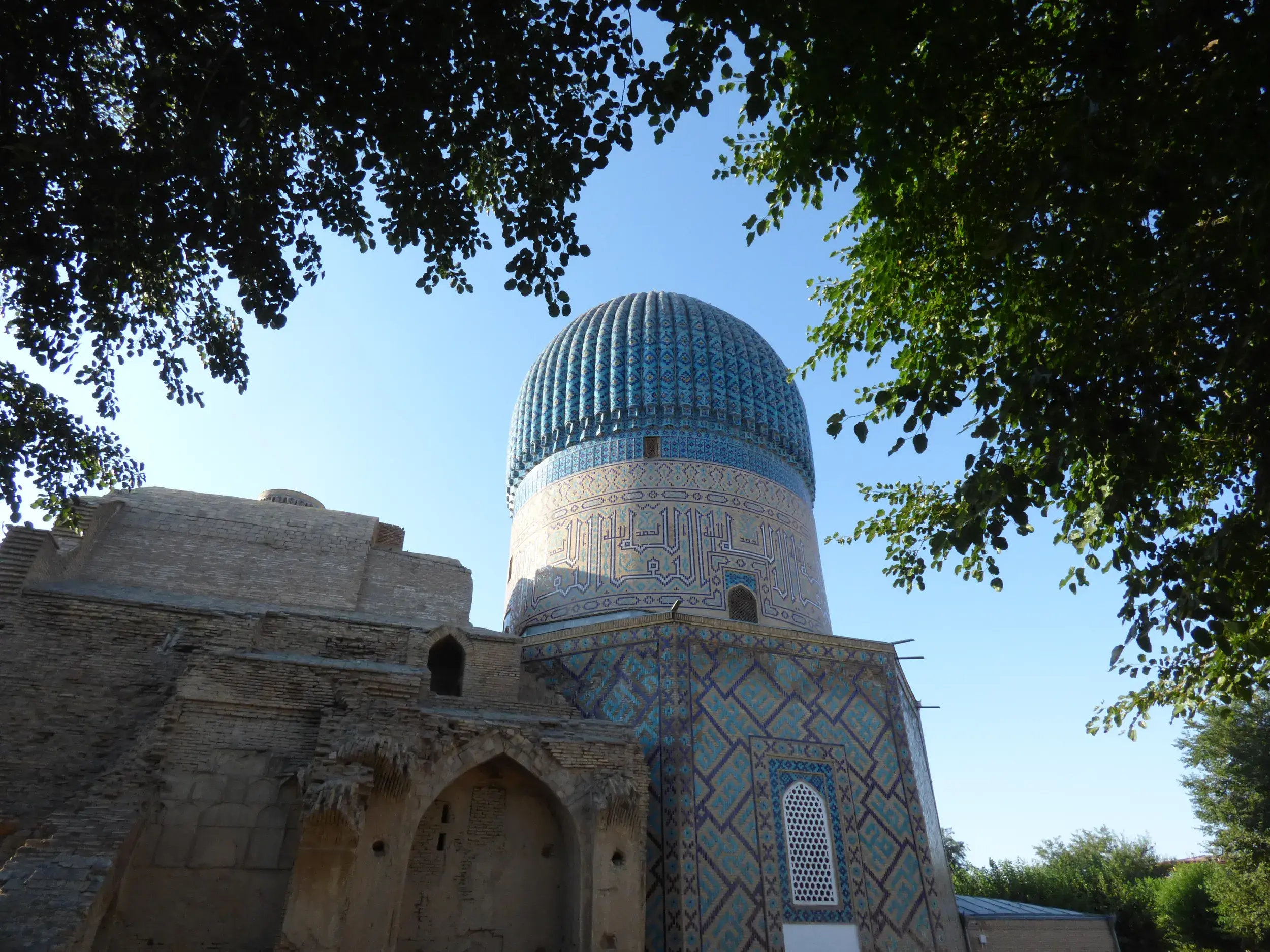 Blick durch grüne Baumkronen auf die prachtvolle, blau gekachelte Kuppel des Gur-Emir-Mausoleums in Samarkand, Usbekistan.