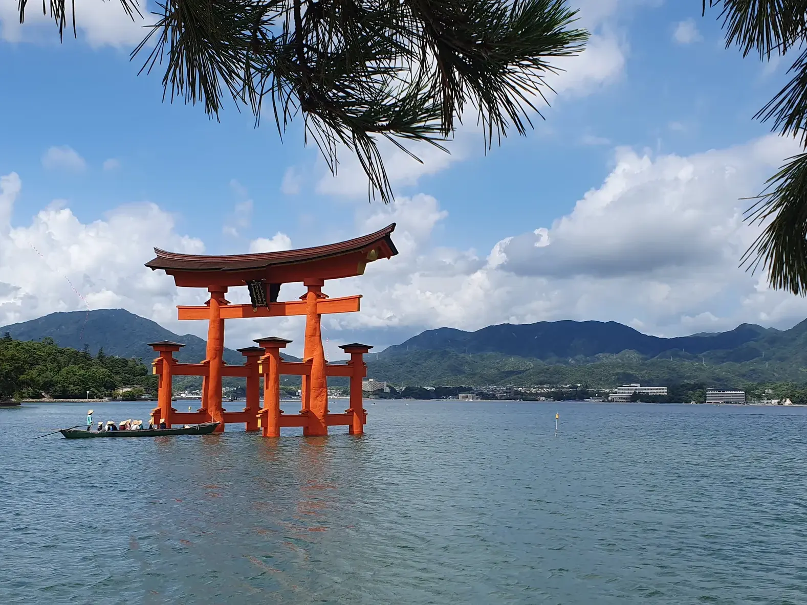 Leuchtend rotes Torii des Itsukushima-Schreins bei Flut im Wasser vor der Küste von Miyajima, im Hintergrund grüne Berge.