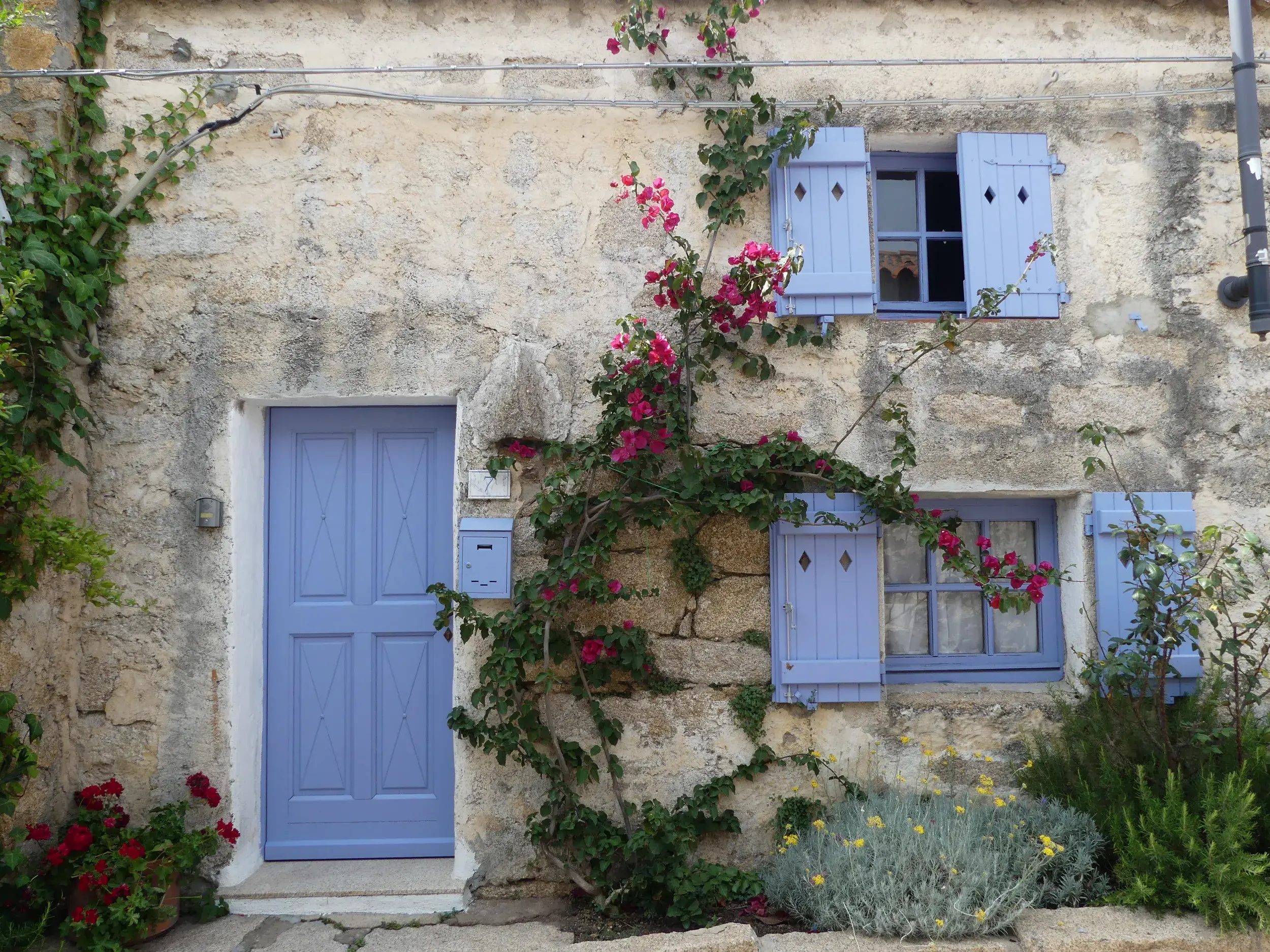 Malerische Steinfassade eines Hauses im Künstlerdorf San Pantaleo auf Sardinien mit blauen Fensterläden und Bougainvillea.