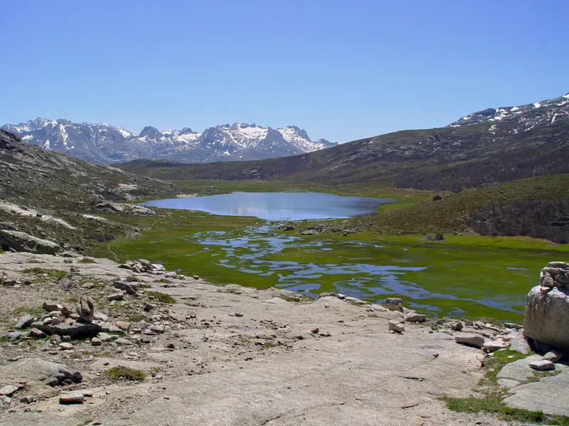 Der Bergsee Lac de Nino auf dem GR20 auf Korsika, eingebettet in grüne Wiesen vor der Kulisse schroffer Berggipfel.