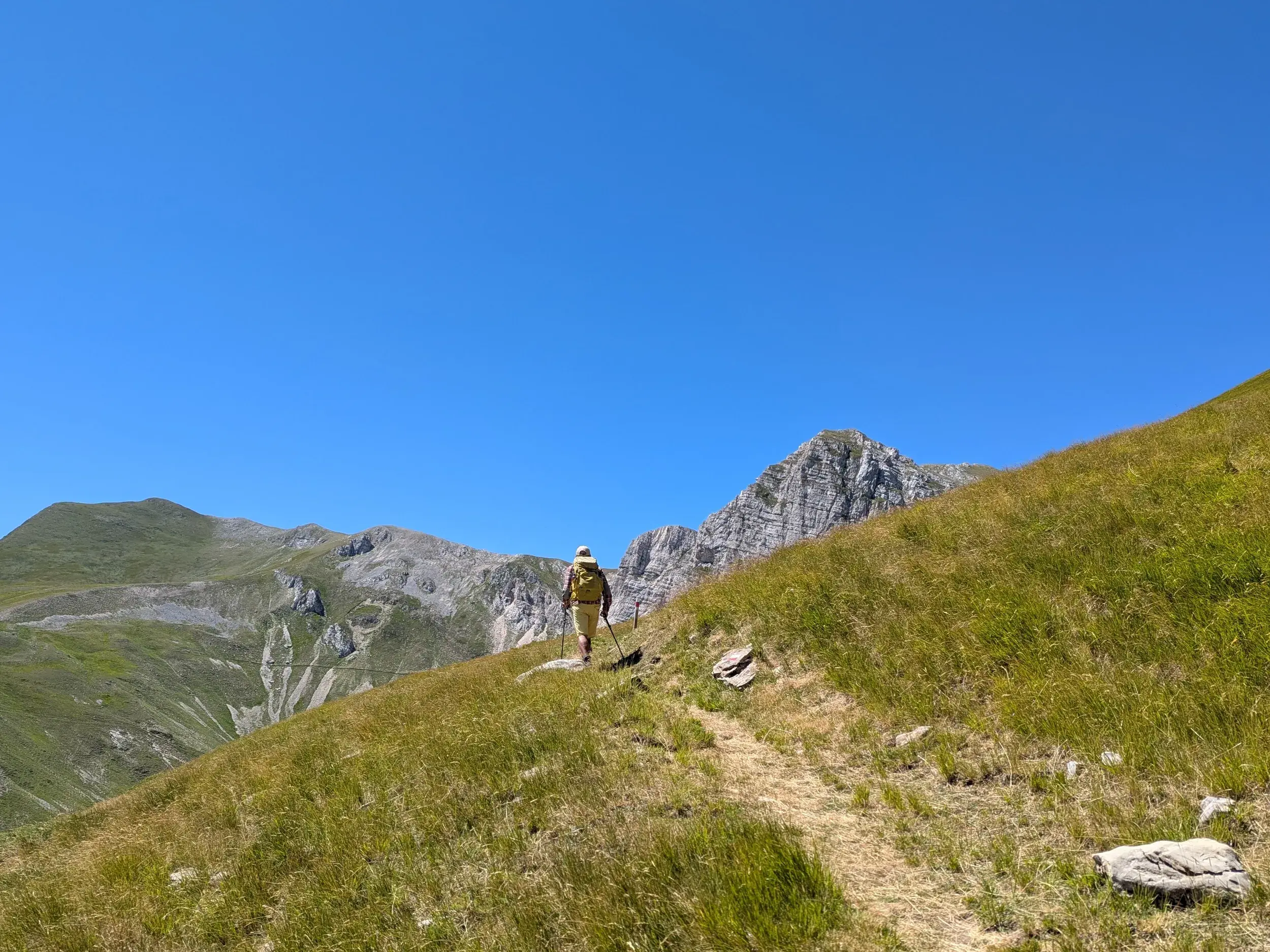 Ein Wanderer mit Rucksack erklimmt einen grünen Berghang in den Sibillinischen Bergen, im Hintergrund ein markanter Felsgipfel.