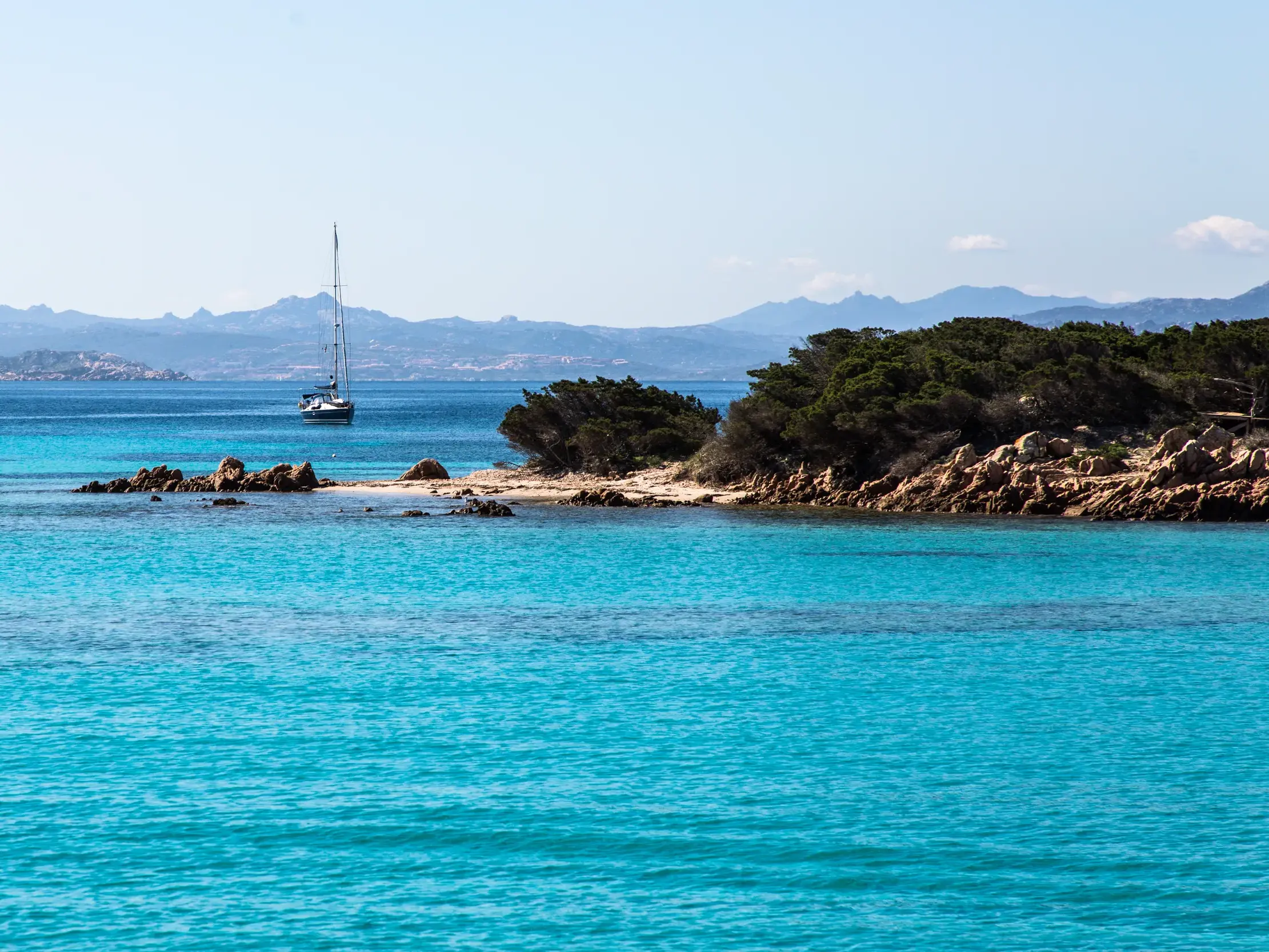 Leuchtend türkisfarbenes Meer an einer felsigen Bucht der Costa Smeralda, im Hintergrund ein Segelboot vor der Bergkulisse Sardiniens.