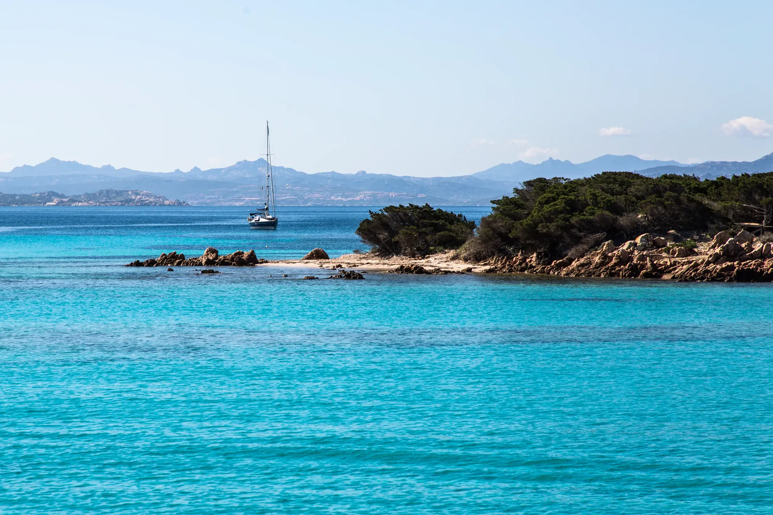 Leuchtend türkisfarbenes Meer an einer felsigen Bucht der Costa Smeralda, im Hintergrund ein Segelboot vor der Bergkulisse Sardiniens.