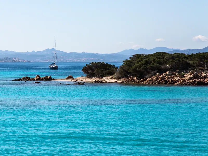 Leuchtend türkisfarbenes Meer an einer felsigen Bucht der Costa Smeralda, im Hintergrund ein Segelboot vor der Bergkulisse Sardiniens.