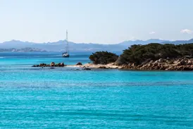 Leuchtend türkisfarbenes Meer an einer felsigen Bucht der Costa Smeralda, im Hintergrund ein Segelboot vor der Bergkulisse Sardiniens.