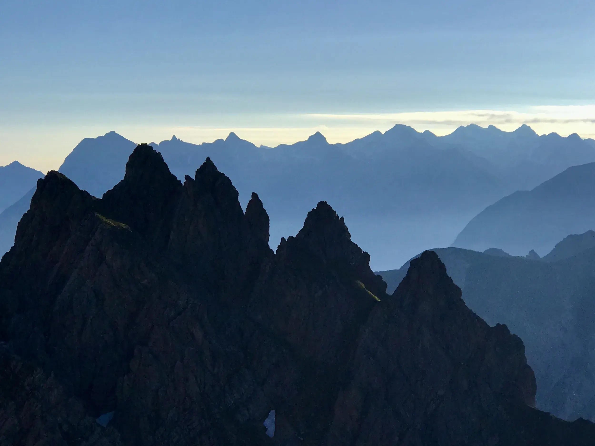 Schroffe Gipfelsilhouetten der Erlspitzgruppe im Karwendel, dahinter gestaffelte Bergketten im blauen Morgendunst.