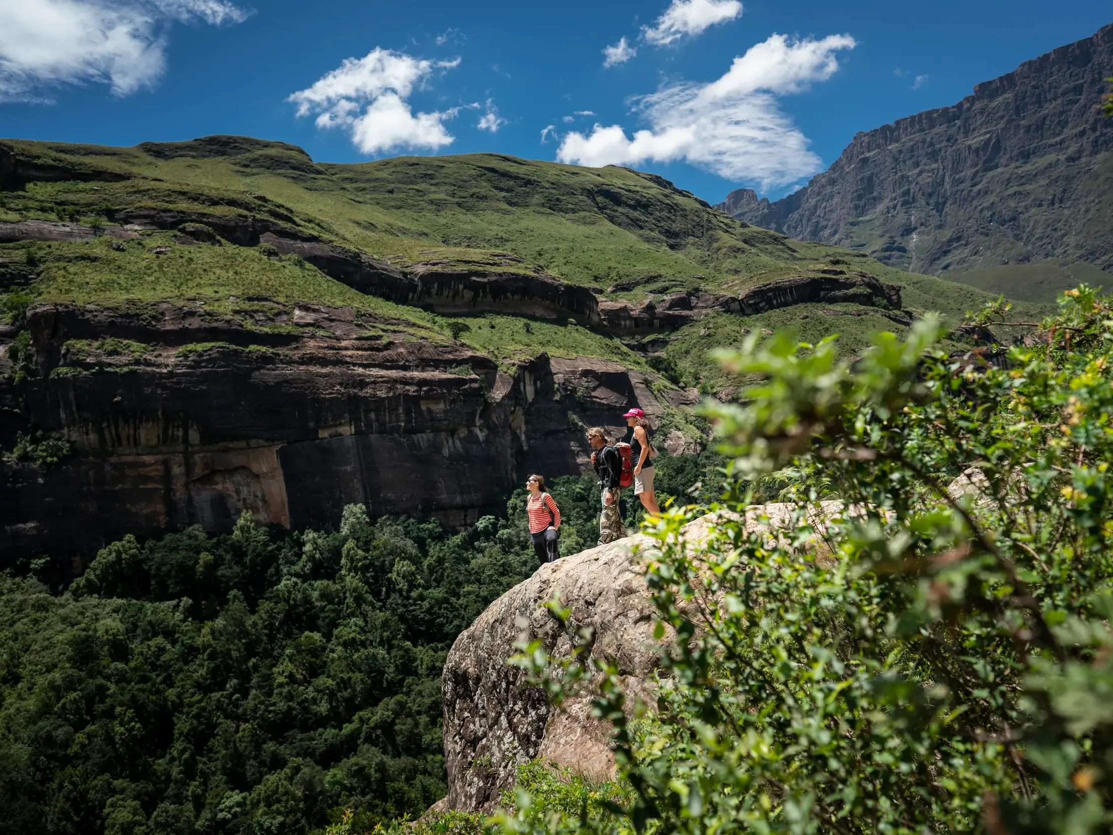 Kleine Wandergruppe auf einem Felsvorsprung genießt die Panoramaaussicht über ein grünes Tal in den Drakensbergen Südafrikas.