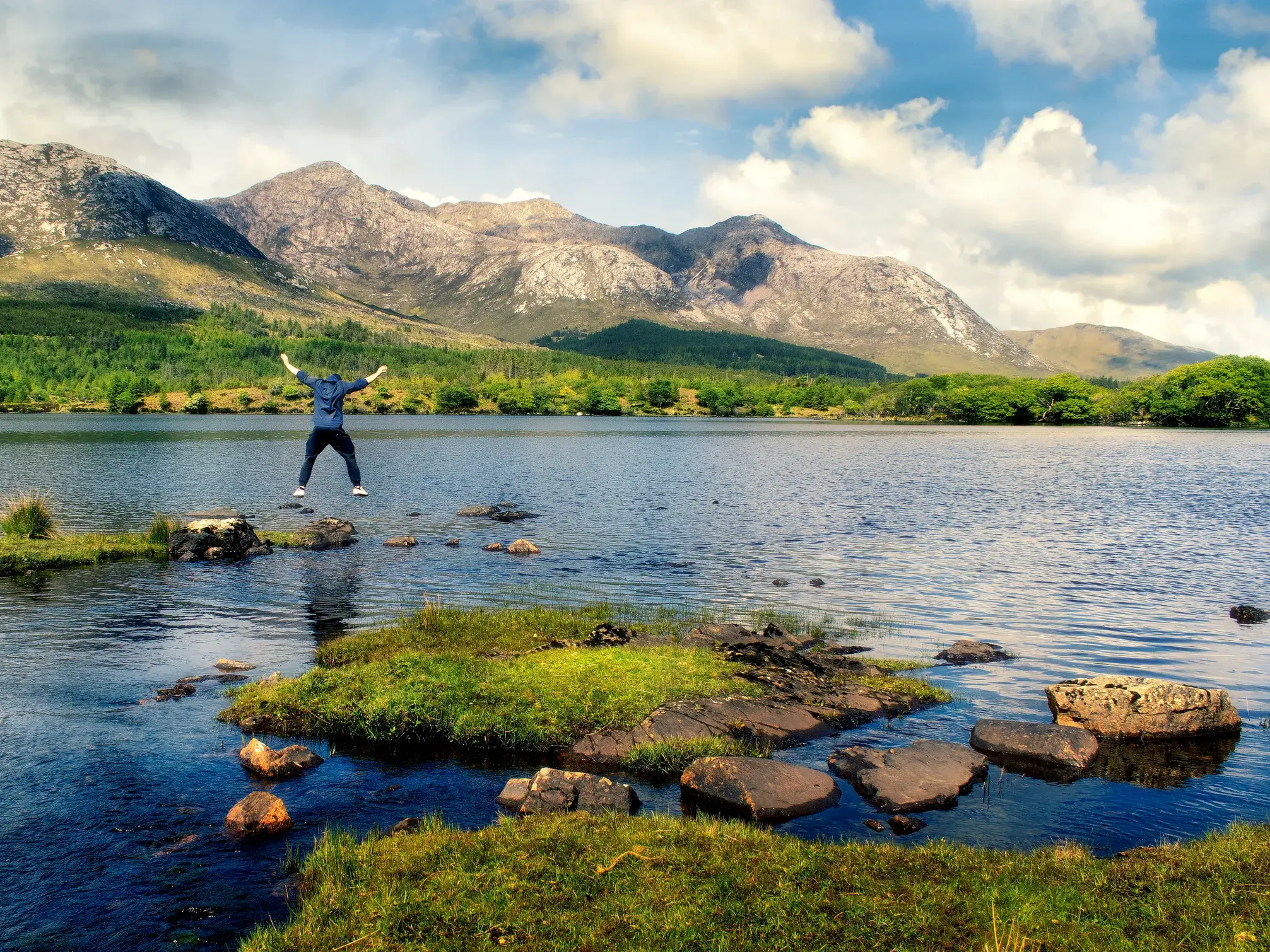 Ein junger Reisender springt euphorisch über Trittsteine am Ufer eines Sees im irischen Connemara-Nationalpark.