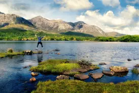 Ein junger Reisender springt euphorisch über Trittsteine am Ufer eines Sees im irischen Connemara-Nationalpark.
