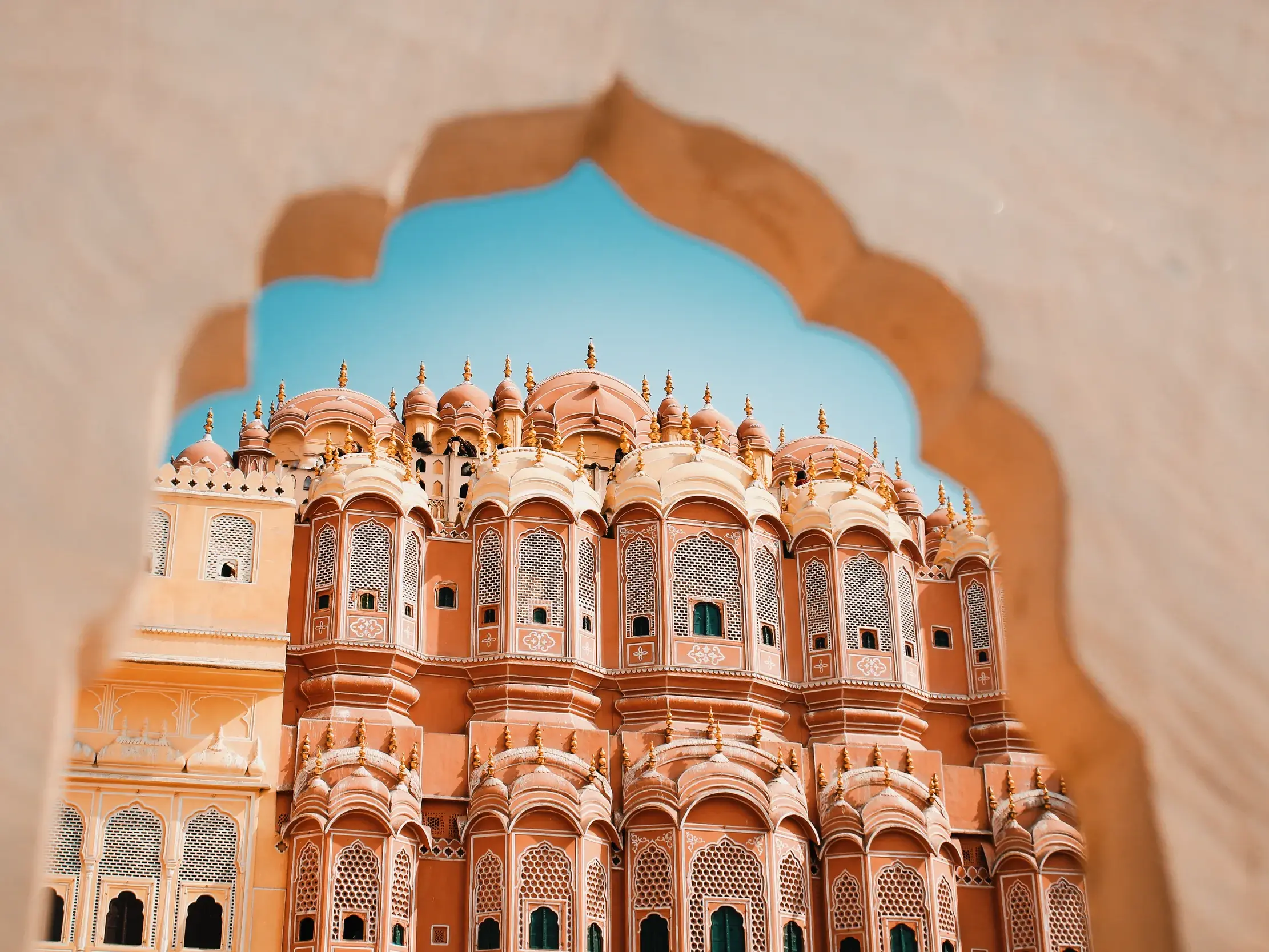 Die kunstvolle, rosafarbene Fassade des Palasts der Winde in Jaipur, gesehen durch einen steinernen Torbogen bei blauem Himmel.