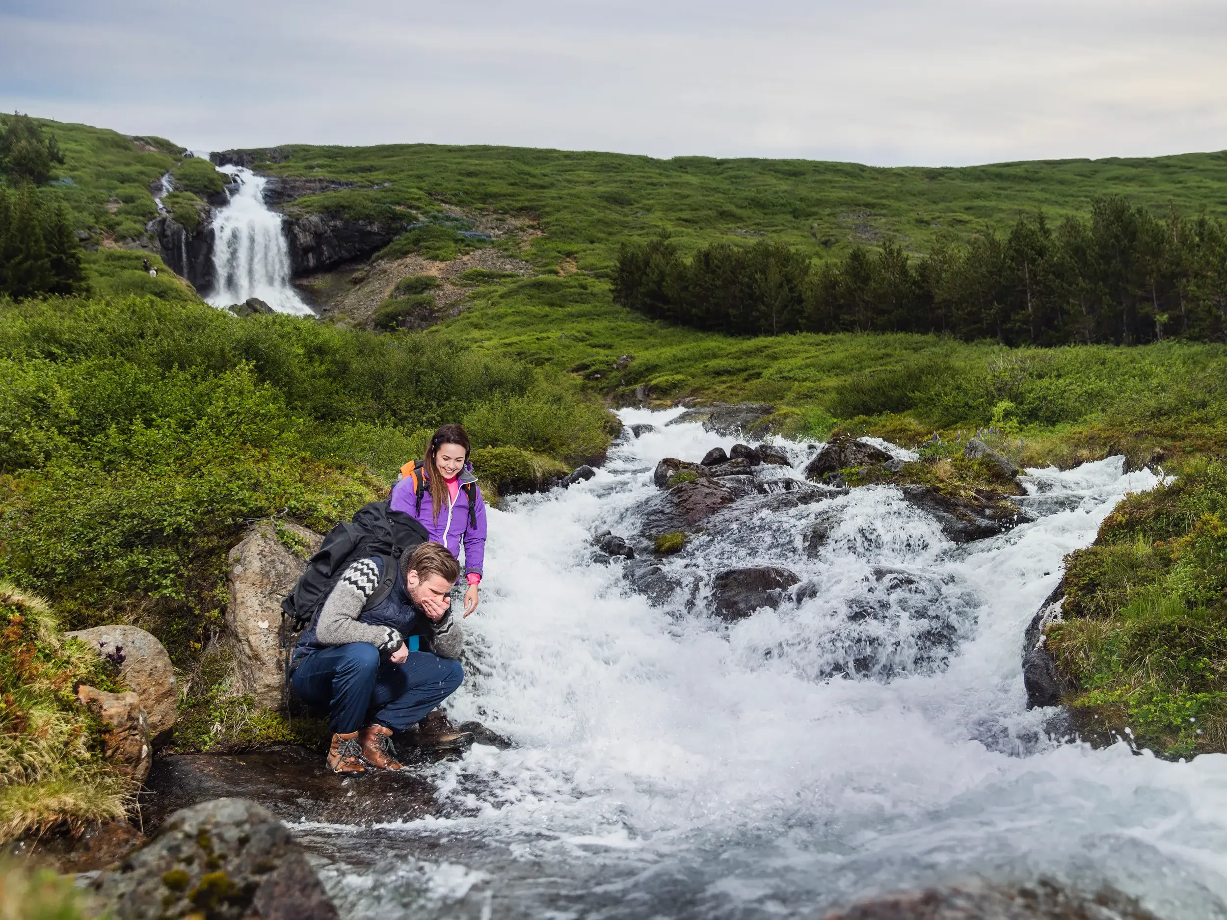 Ein Wanderer erfrischt sich an einem reißenden Bach in der grünen Hügellandschaft Islands, im Hintergrund ein Wasserfall.