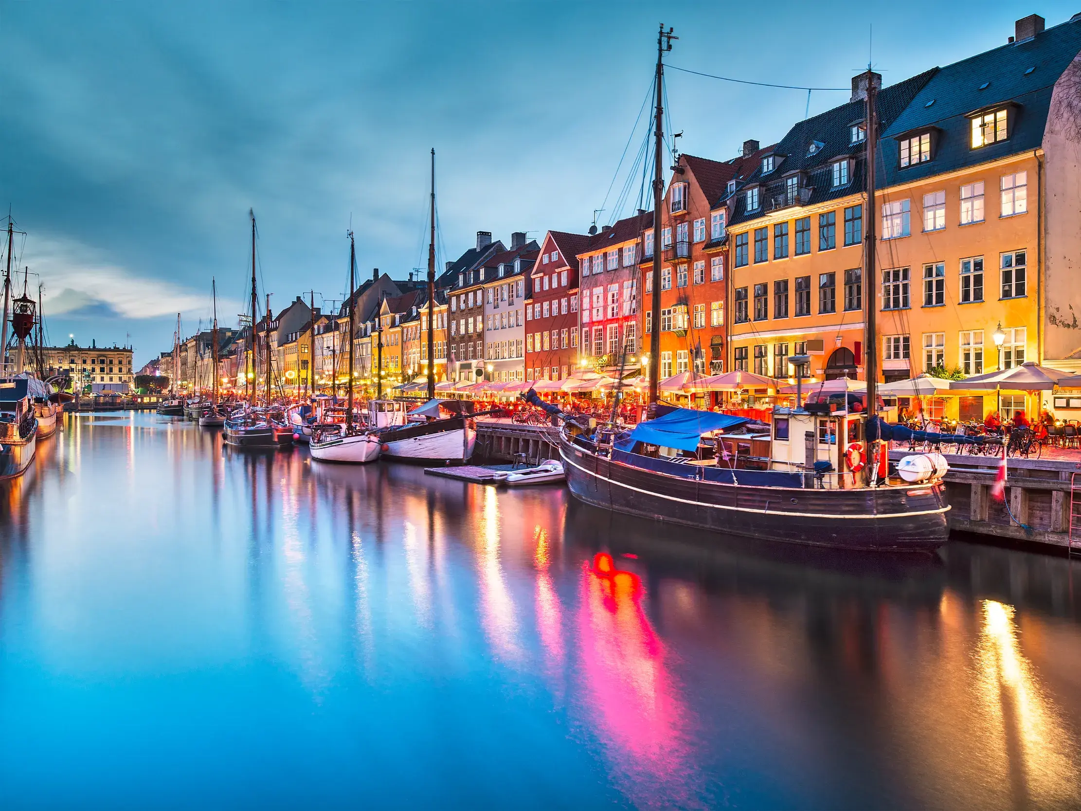 Bunte, beleuchtete Giebelhäuser und historische Schiffe am Nyhavn in Kopenhagen, deren Lichter sich im Kanal spiegeln.