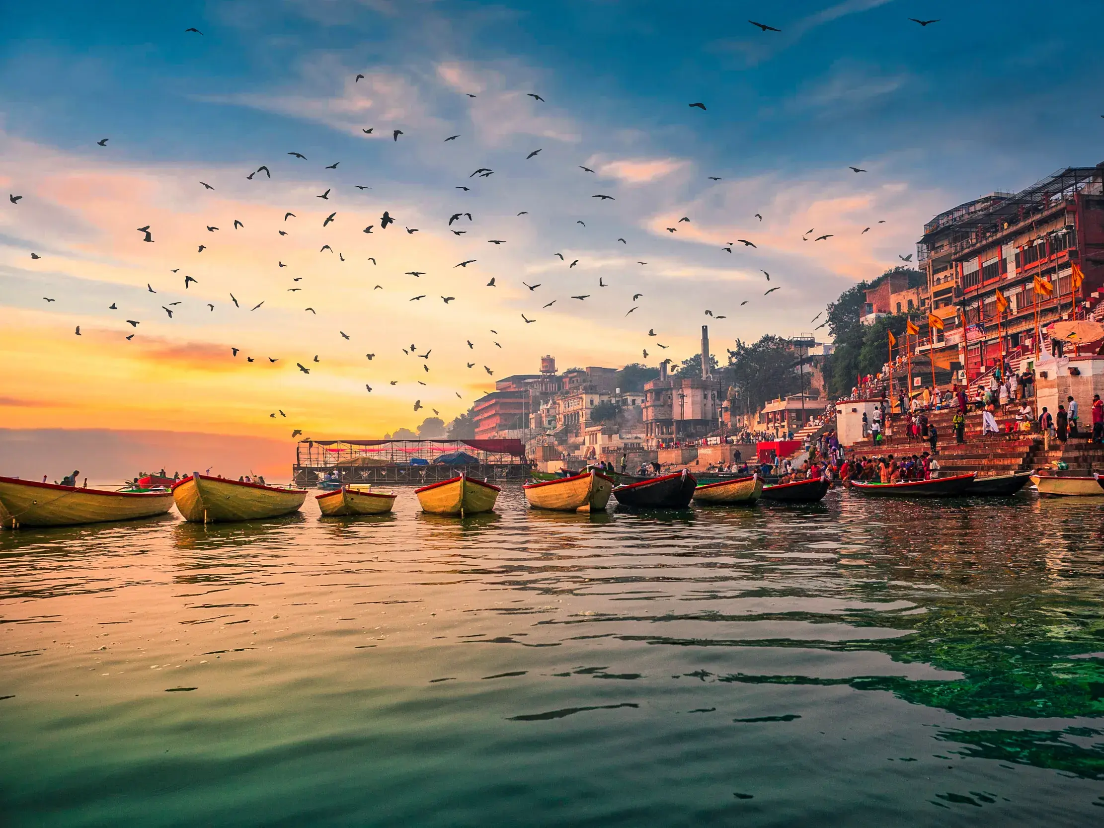 Holzboote treiben bei Sonnenaufgang auf dem Ganges in Varanasi, während Vögel über den belebten Ghats am Ufer fliegen.