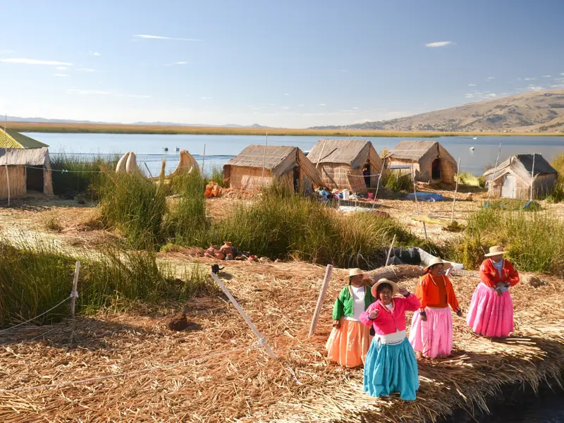 Vier Frauen des Uros-Volkes in bunten Trachten auf einer schwimmenden Schilfinsel mit Hütten am Titicacasee in Peru.