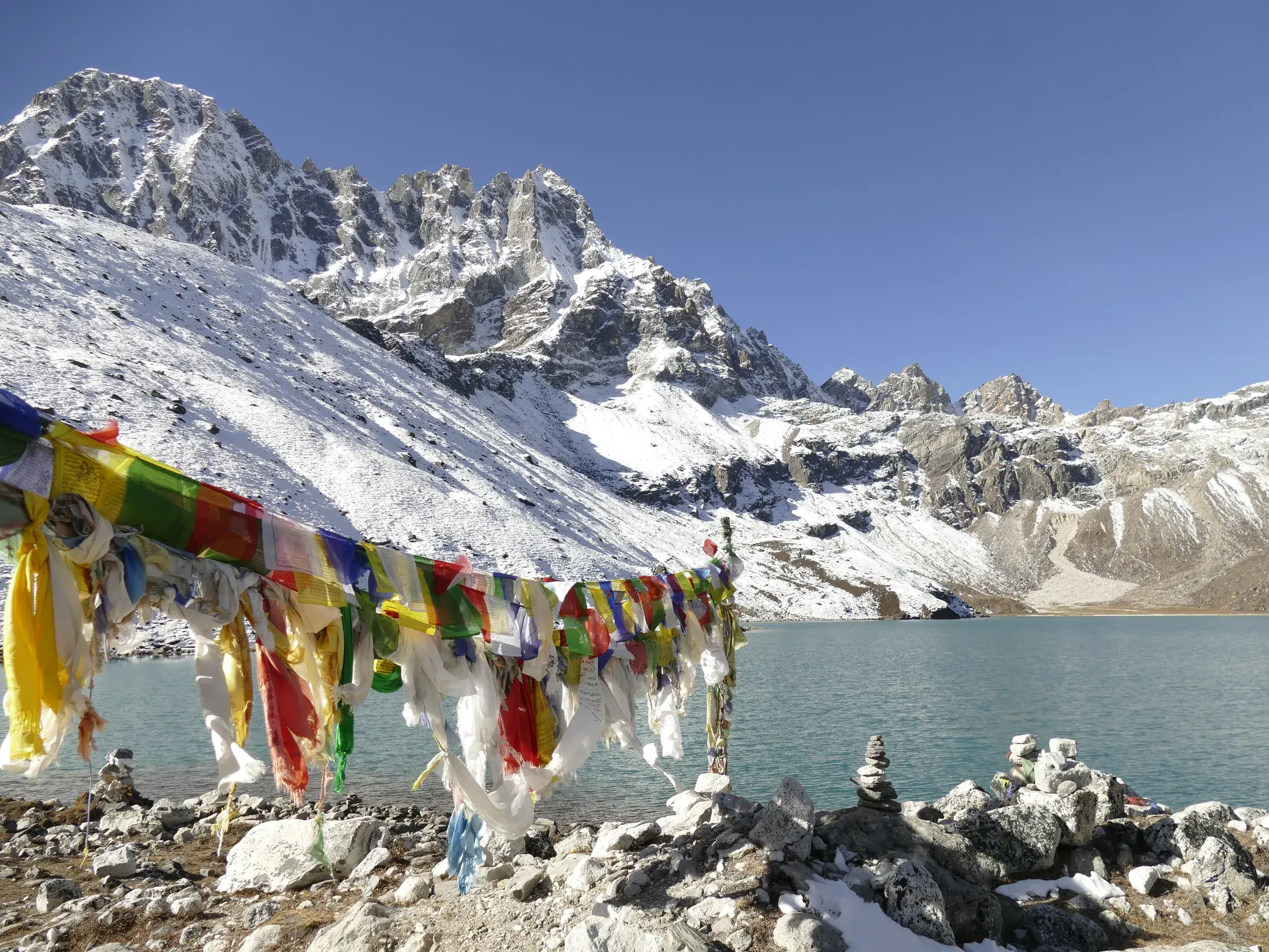 Bunte Gebetsfahnen und Steinmännchen am türkisfarbenen Gokyo-See mit Blick auf die schneebedeckten Gipfel der Khumbu-Region.