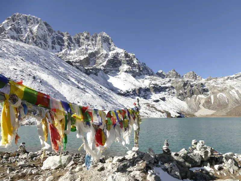 Bunte Gebetsfahnen und Steinmännchen am türkisfarbenen Gokyo-See mit Blick auf die schneebedeckten Gipfel der Khumbu-Region.