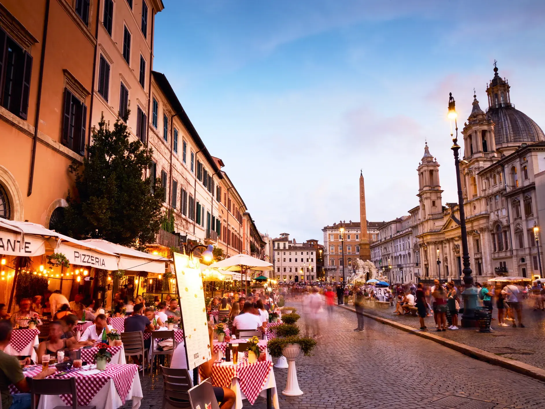 Gäste einer belebten Trattoria auf der Piazza Navona in Rom, im Hintergrund der berühmte Vier-Ströme-Brunnen bei Dämmerung.