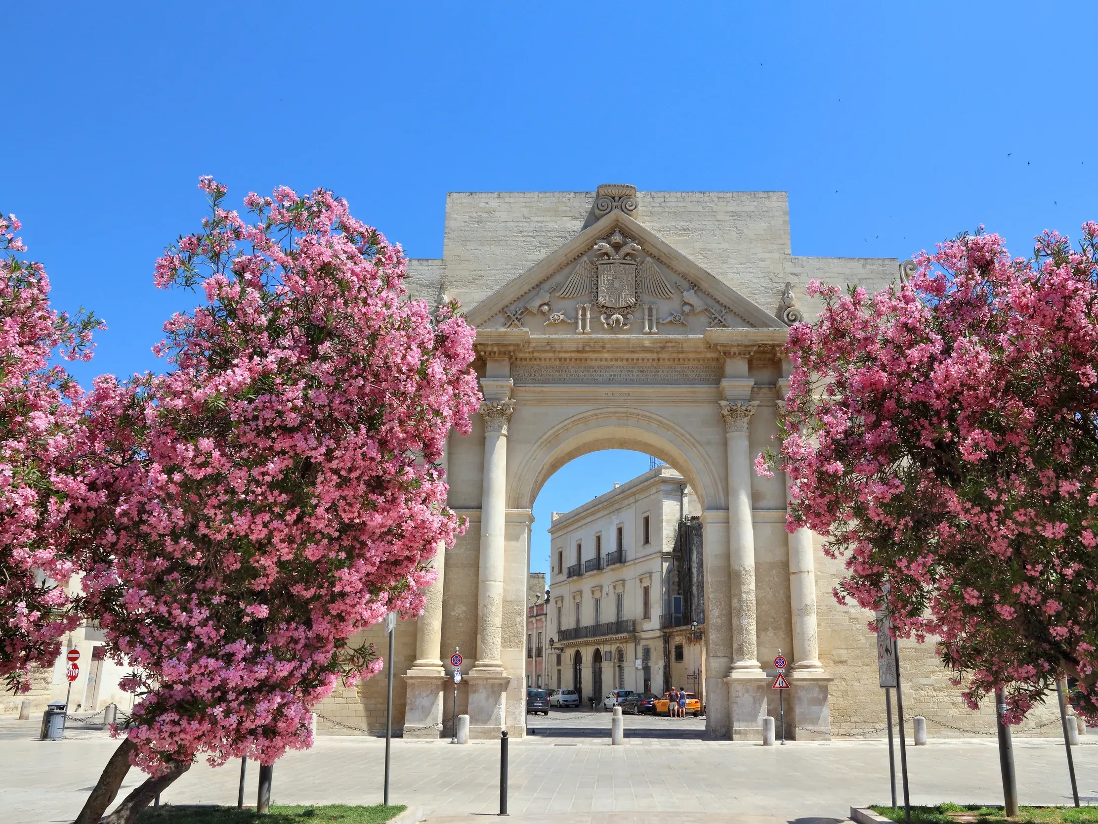 Historisches Stadttor in der Barockstadt Lecce, eingerahmt von rosa blühenden Oleandern unter strahlend blauem Himmel.