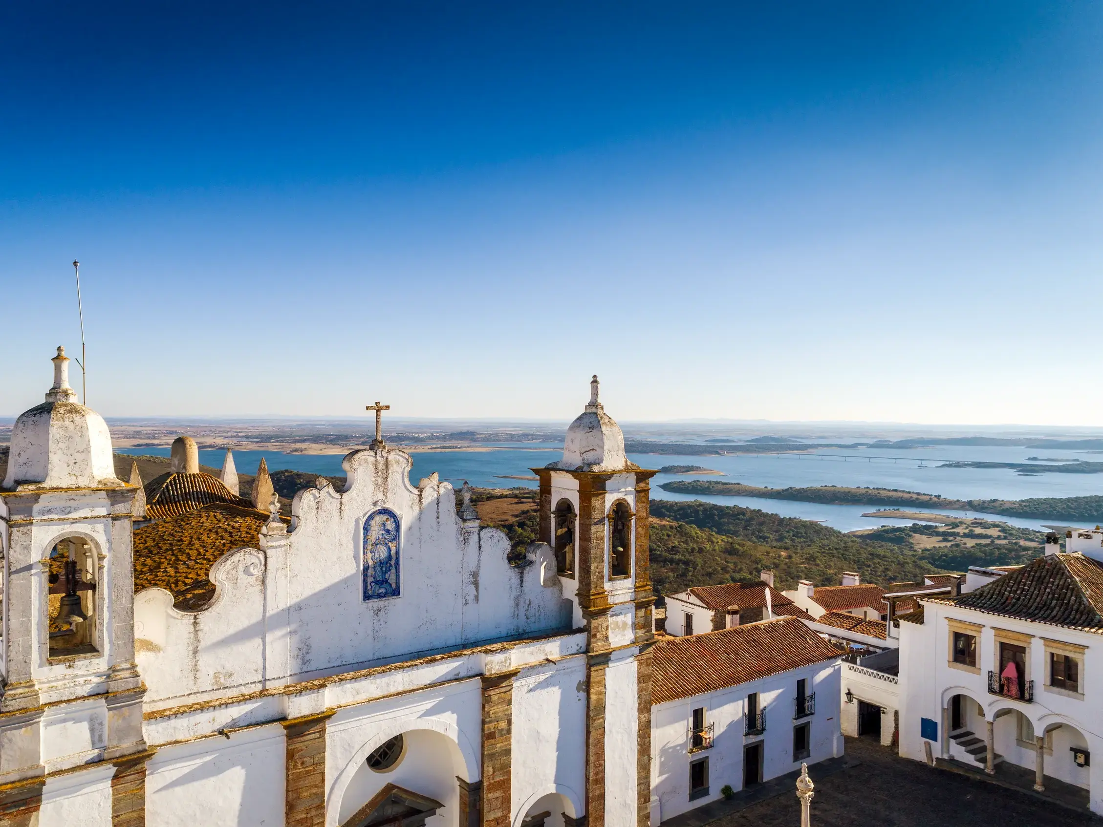 Malerische, weiße Kirche auf einem Hügel im Alentejo mit weitem Blick über eine sonnige Seenlandschaft und die hügelige Ebene.