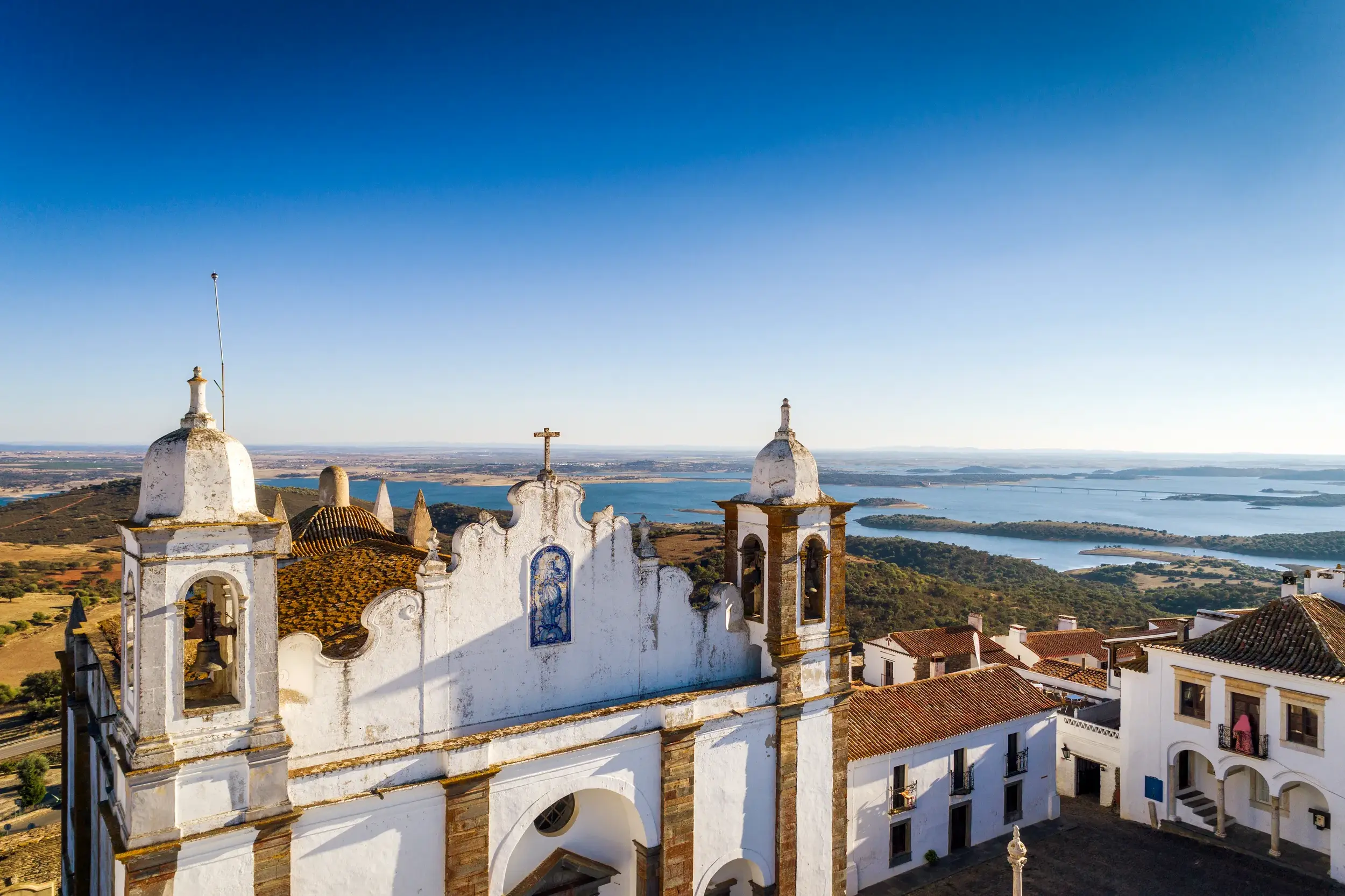 Malerische, weiße Kirche auf einem Hügel im Alentejo mit weitem Blick über eine sonnige Seenlandschaft und die hügelige Ebene.