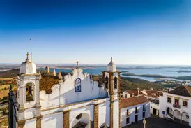 Malerische, weiße Kirche auf einem Hügel im Alentejo mit weitem Blick über eine sonnige Seenlandschaft und die hügelige Ebene.