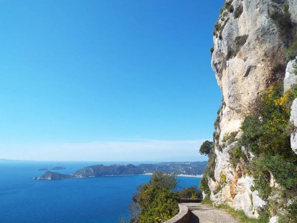 Schmaler Wanderpfad an einer Felsklippe mit Blick auf die Buchten von Paleokastritsa und das tiefblaue Ionische Meer auf Korfu.