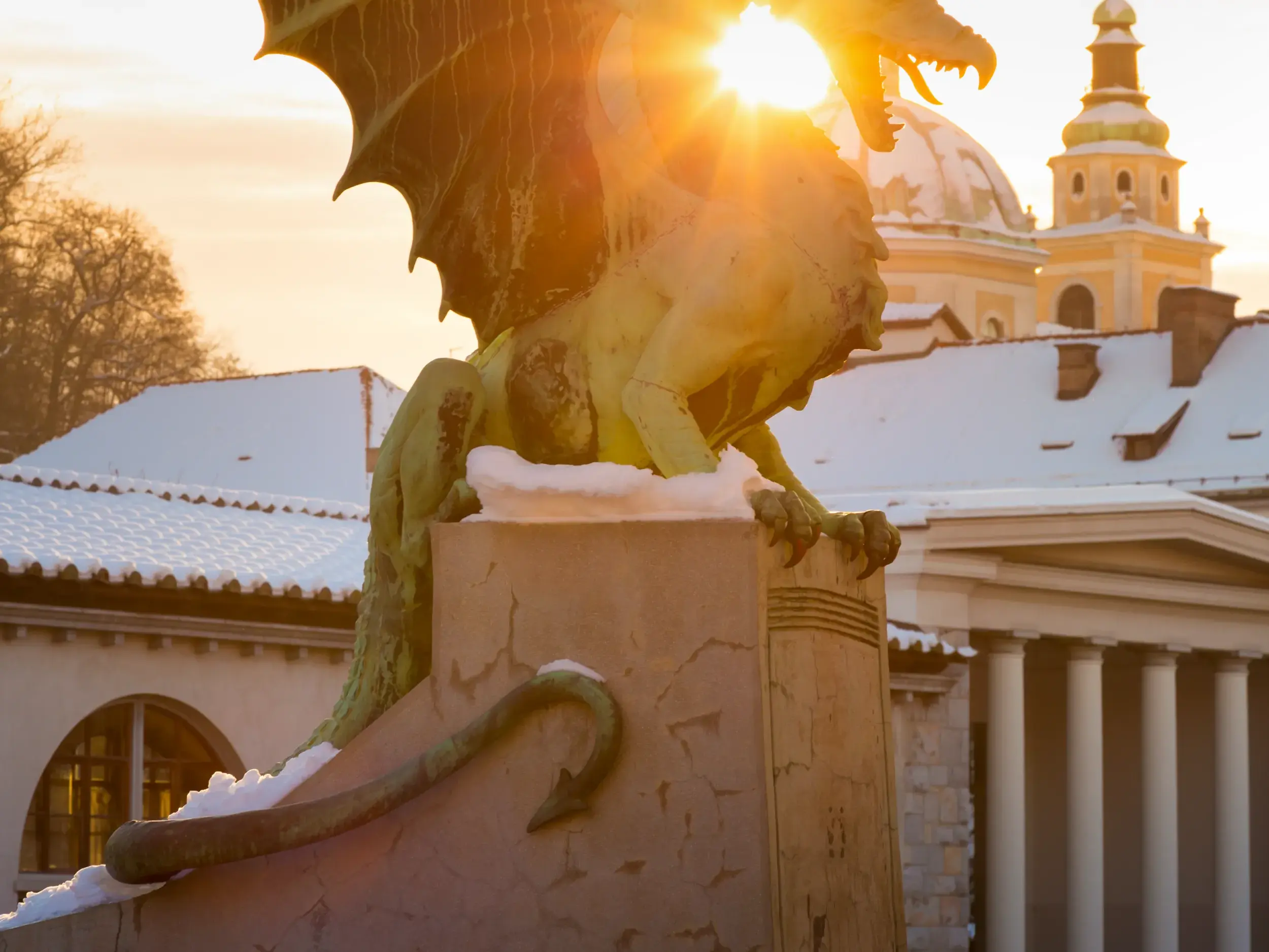 Berühmte Drachenstatue in Ljubljana, im warmen Licht der Wintersonne über schneebedeckten Dächern der Altstadt.