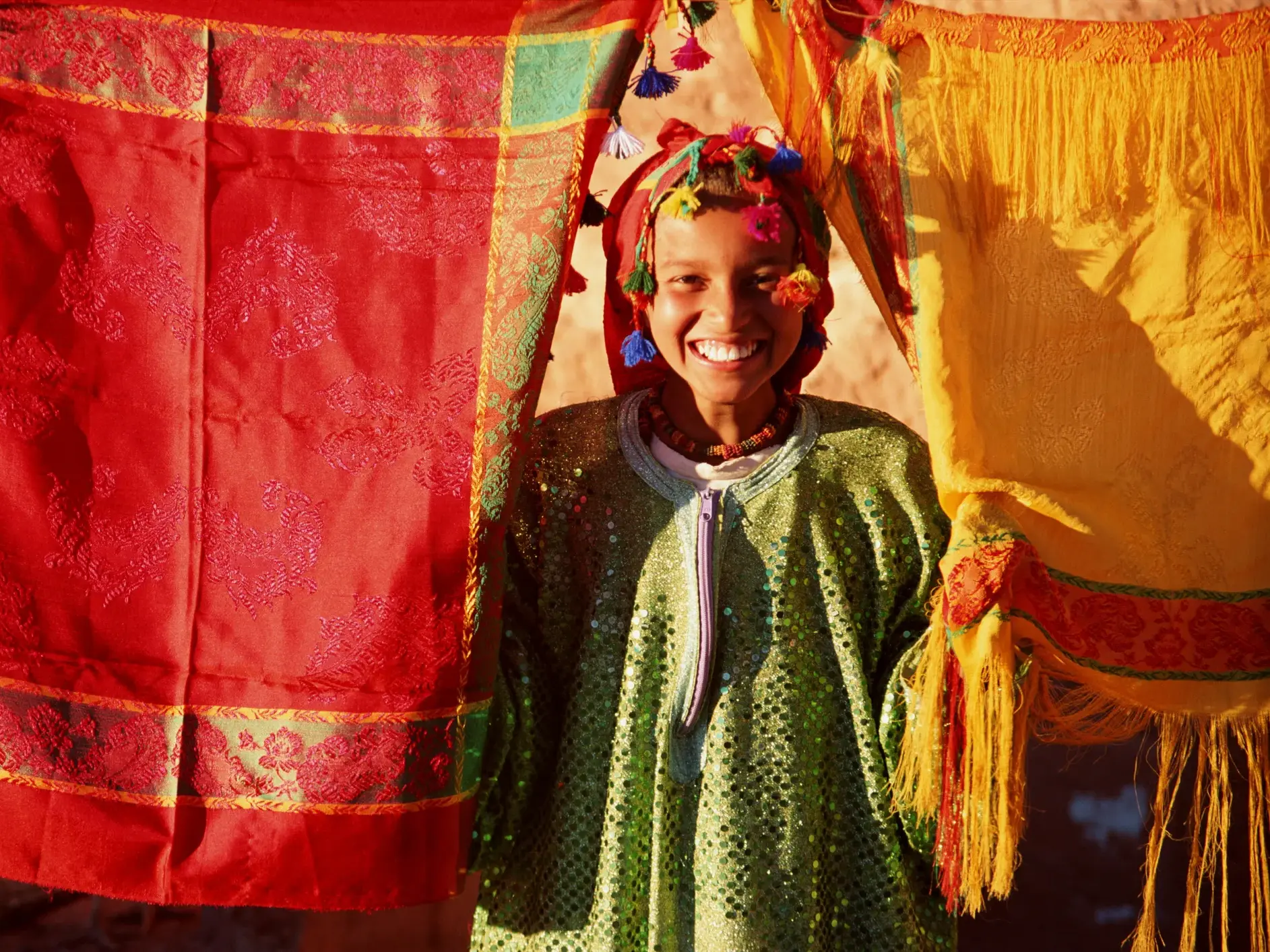 Ein junges Mädchen in traditioneller Kleidung lächelt fröhlich zwischen farbenfrohen Textilien auf einem Souk in Marrakesch.