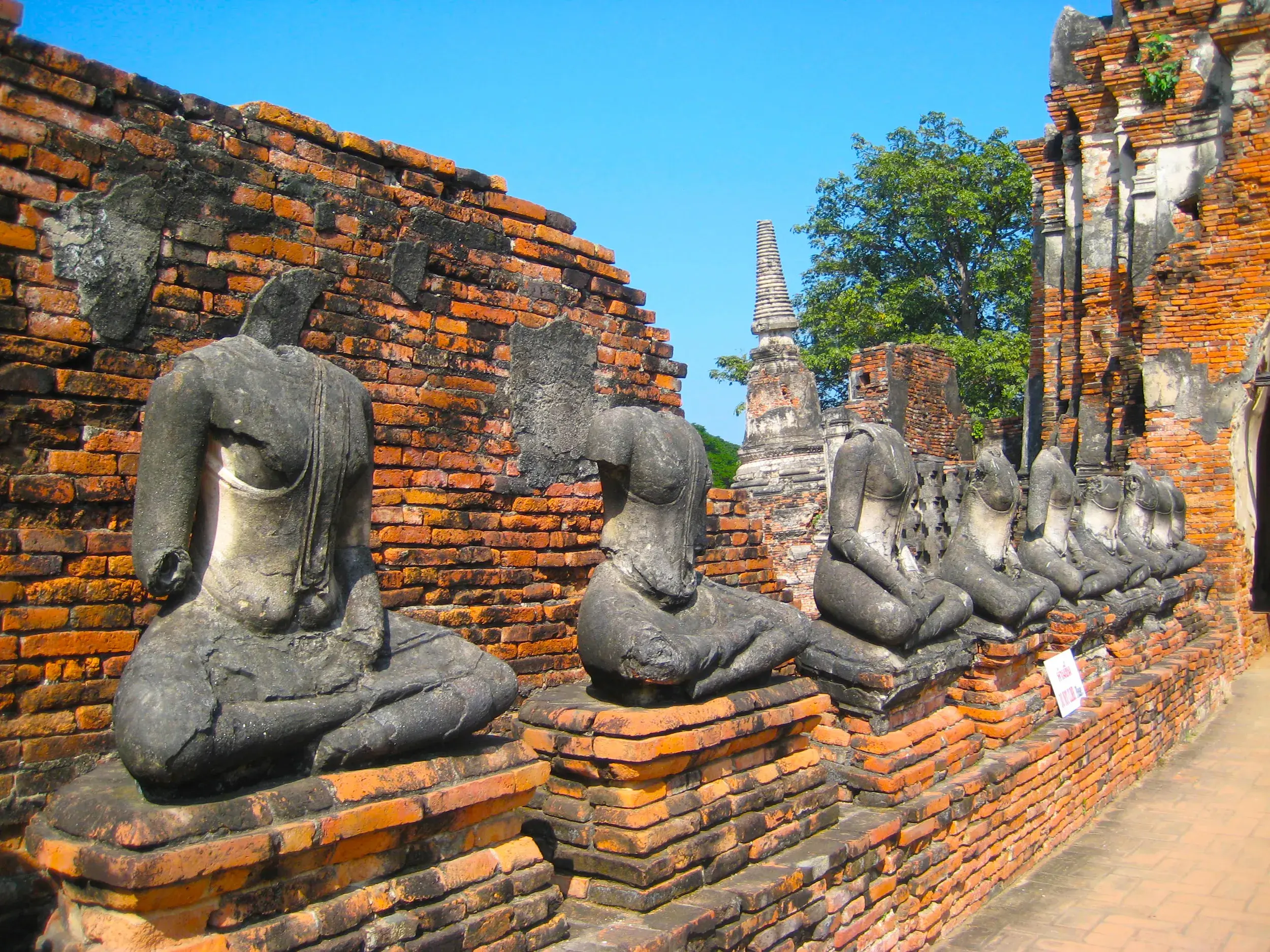 Alte, kopflose Buddha-Statuen sitzen in einer Reihe vor einer roten Ziegelmauer in der Tempelanlage von Ayutthaya.