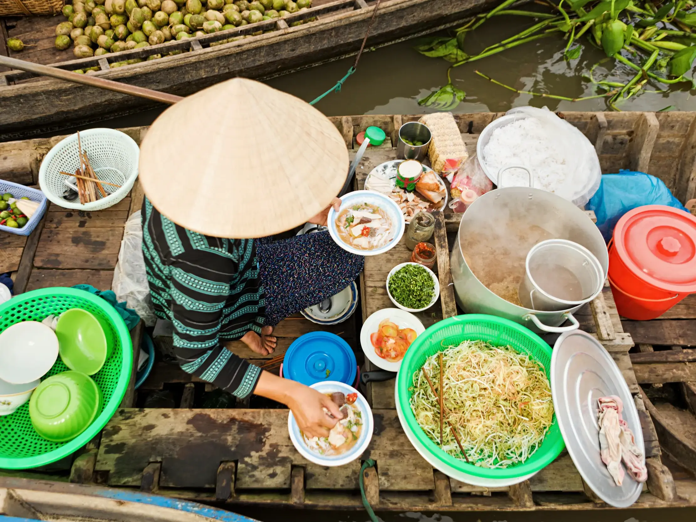 Vietnamesische Händlerin mit Kegelhut serviert Nudelsuppe aus ihrer Bootsküche auf dem schwimmenden Markt im Mekongdelta.