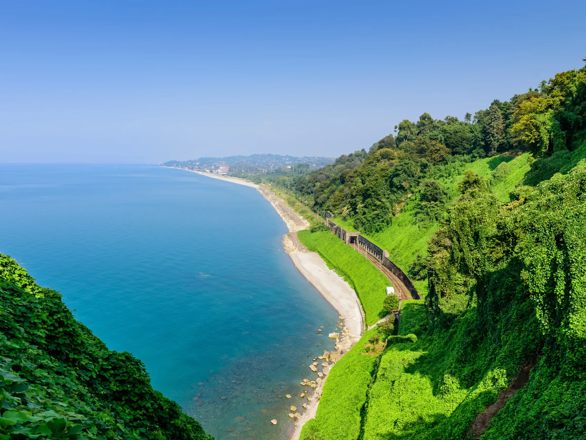 Blick vom Botanischen Garten Batumi auf die üppig grüne Schwarzmeerküste, an der ein Zug entlang der Klippen fährt.
