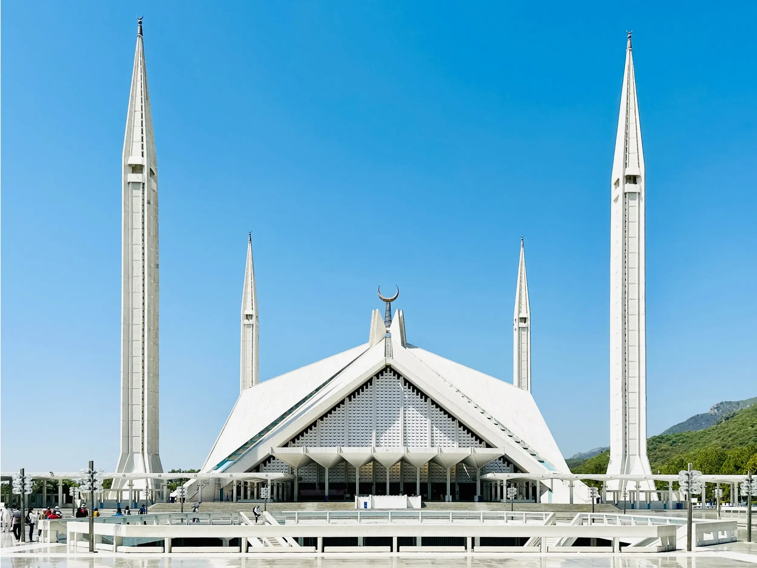 Die moderne Architektur der weißen Faisal-Moschee in Islamabad mit ihren spitzen Minaretten vor den grünen Margalla Hills.